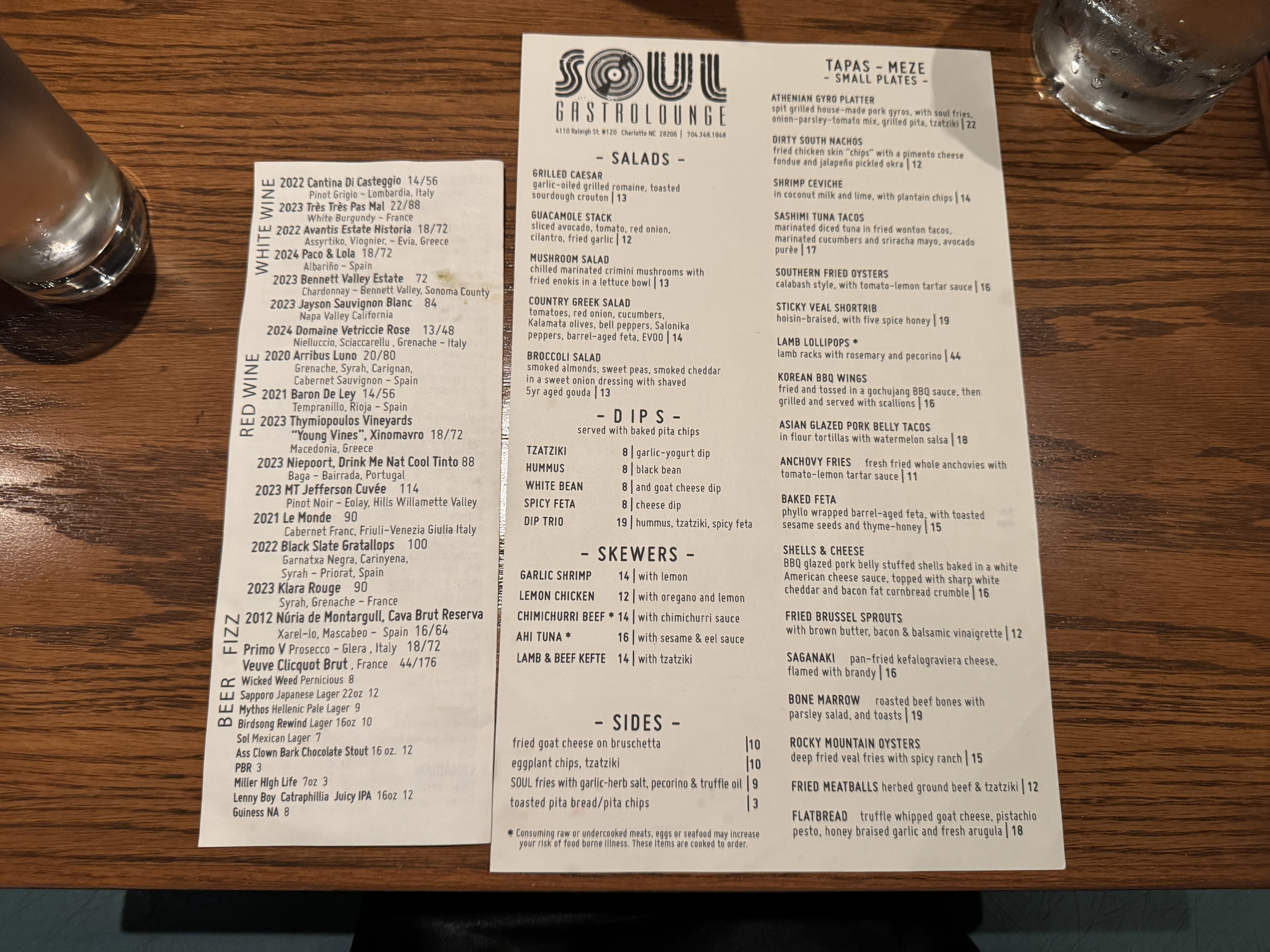 Menu from Soul Gastrolounge featuring salads, dips, skewers, sides, and tapas with prices; sections for white and red wines, beer, and fizz; menu on a wooden table with two glasses of water.