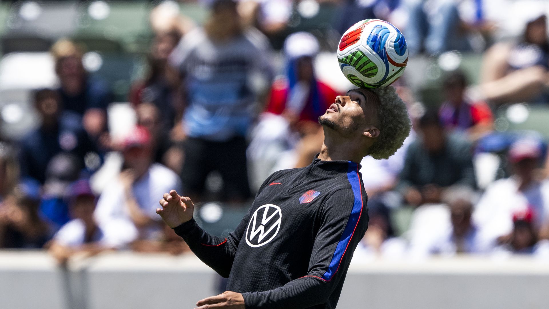 U.S. Men's National Team player Antonee Robinson balances a green, blue, red and white soccer ball on his head. People behind him are blurred out.