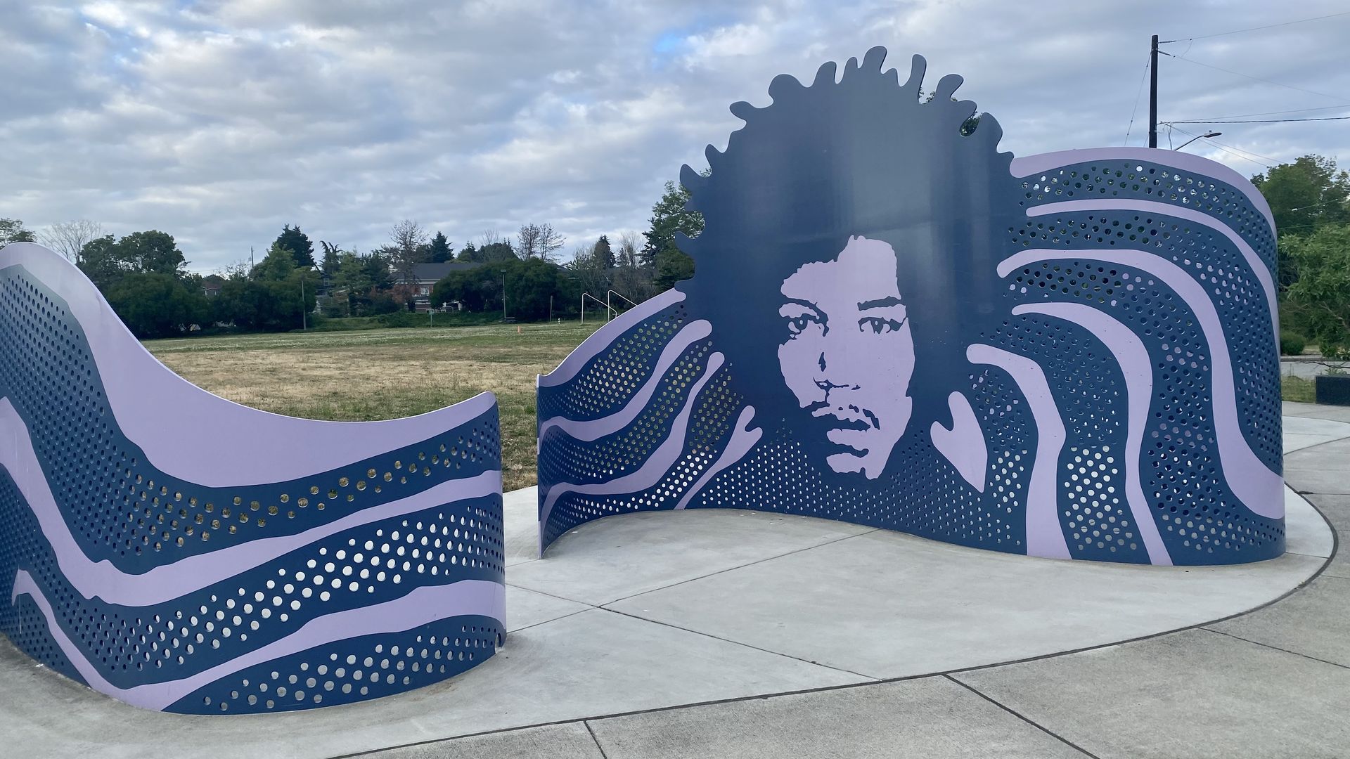 A purple metal sculpture with the face of Jimi Hendrix featured prominently, with clouds above, grass and trees in background.