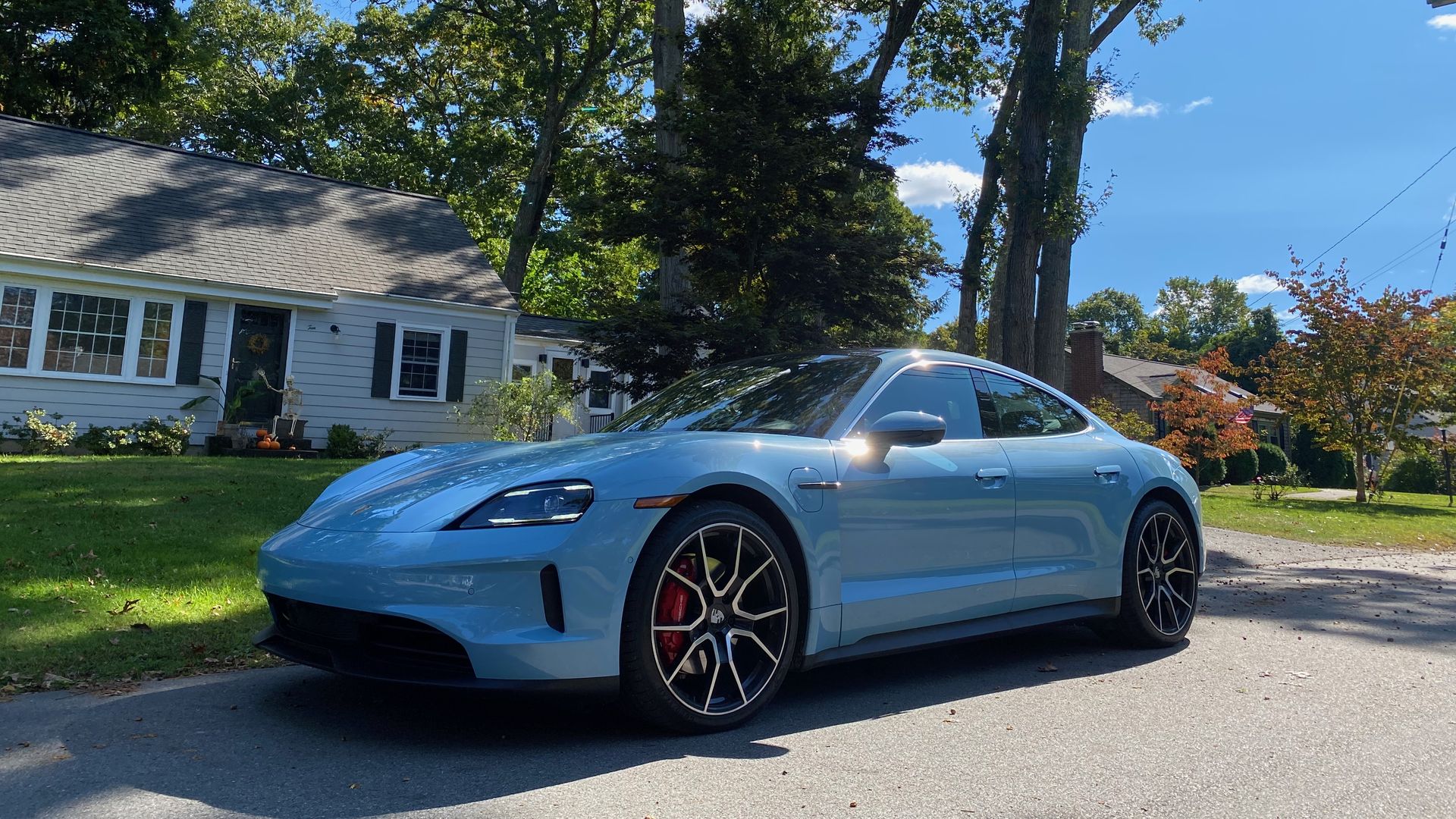 A photo of an electric blue Porsche Taycan four-door EV, parked on a suburban street in front of a green lawn, some trees, and a house.