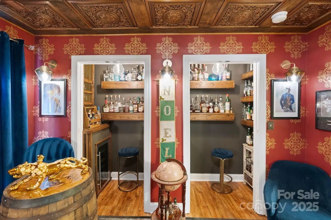 Room with red patterned wallpaper, carved wooden ceiling, two small bar areas with stools, shelves of liquor bottles, deep blue velvet chairs, a wooden barrel with a golden panther sculpture, and a globe stand.