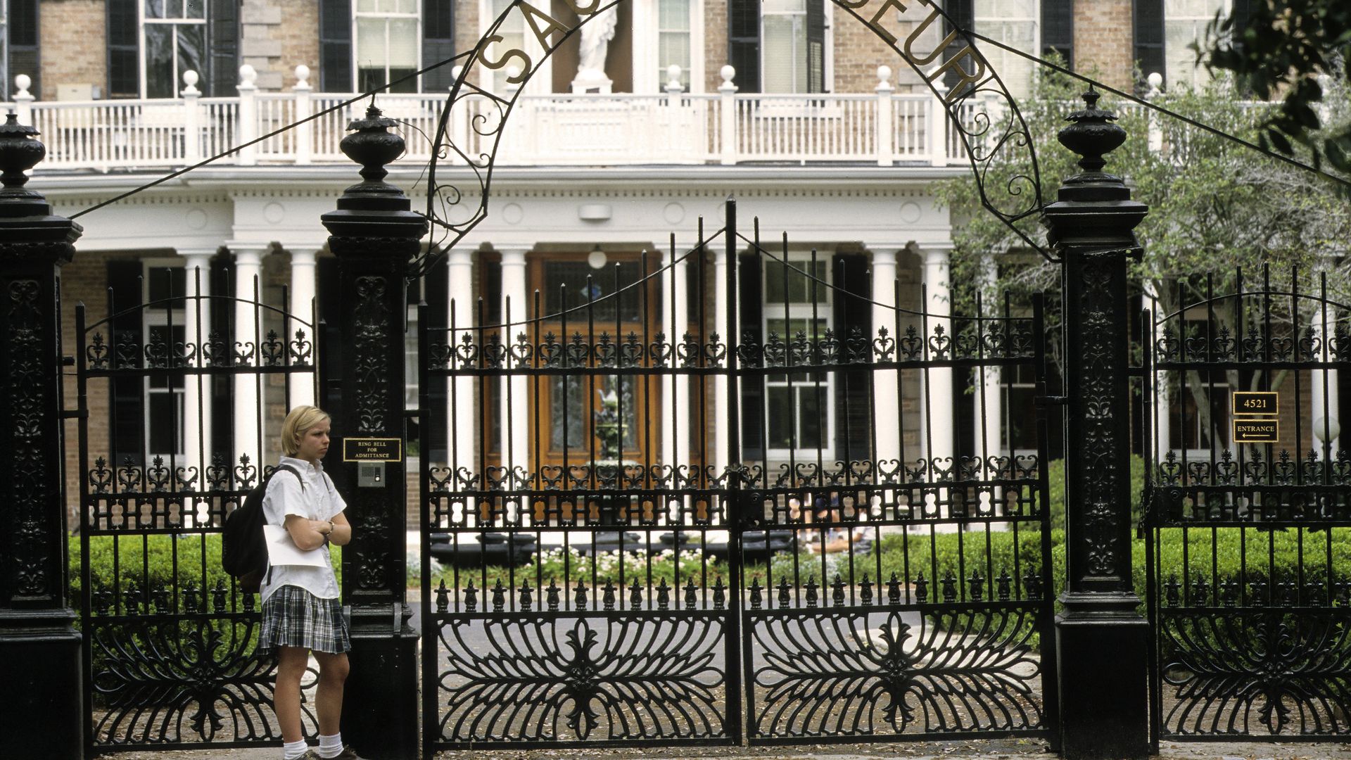 The photo shows a student in uniform outside the tall metal gate at Sacred Heart in New Orleans. The school building is visible in the background.