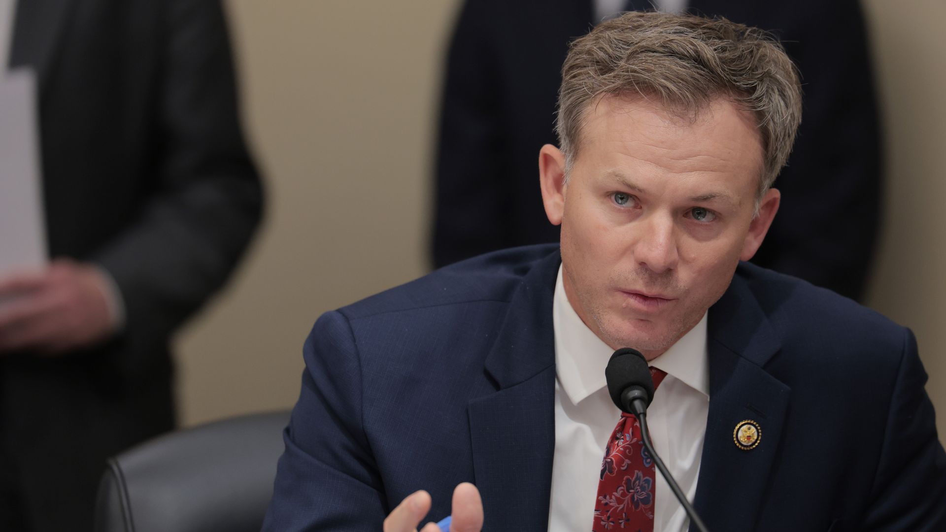 Man with short light brown hair speaking into a microphone, wearing a navy suit and red floral tie, seated behind a desk labeled "Mr. Moore (UT)".