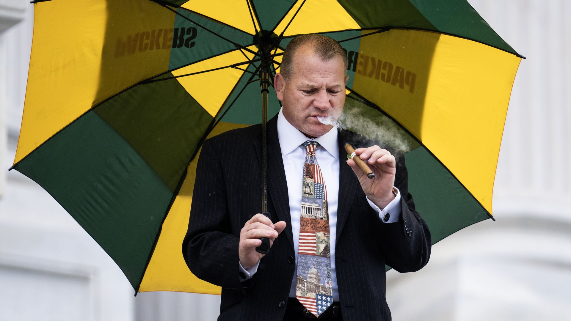 Rep. Troy Nehls, carrying a yellow and green Green Bay Packers umbrella and smoking a cigar outside the Capitol while wearing a black suit, light blue shirt and tie with American flags, eagles, U.S. monuments and the Constitution on it.