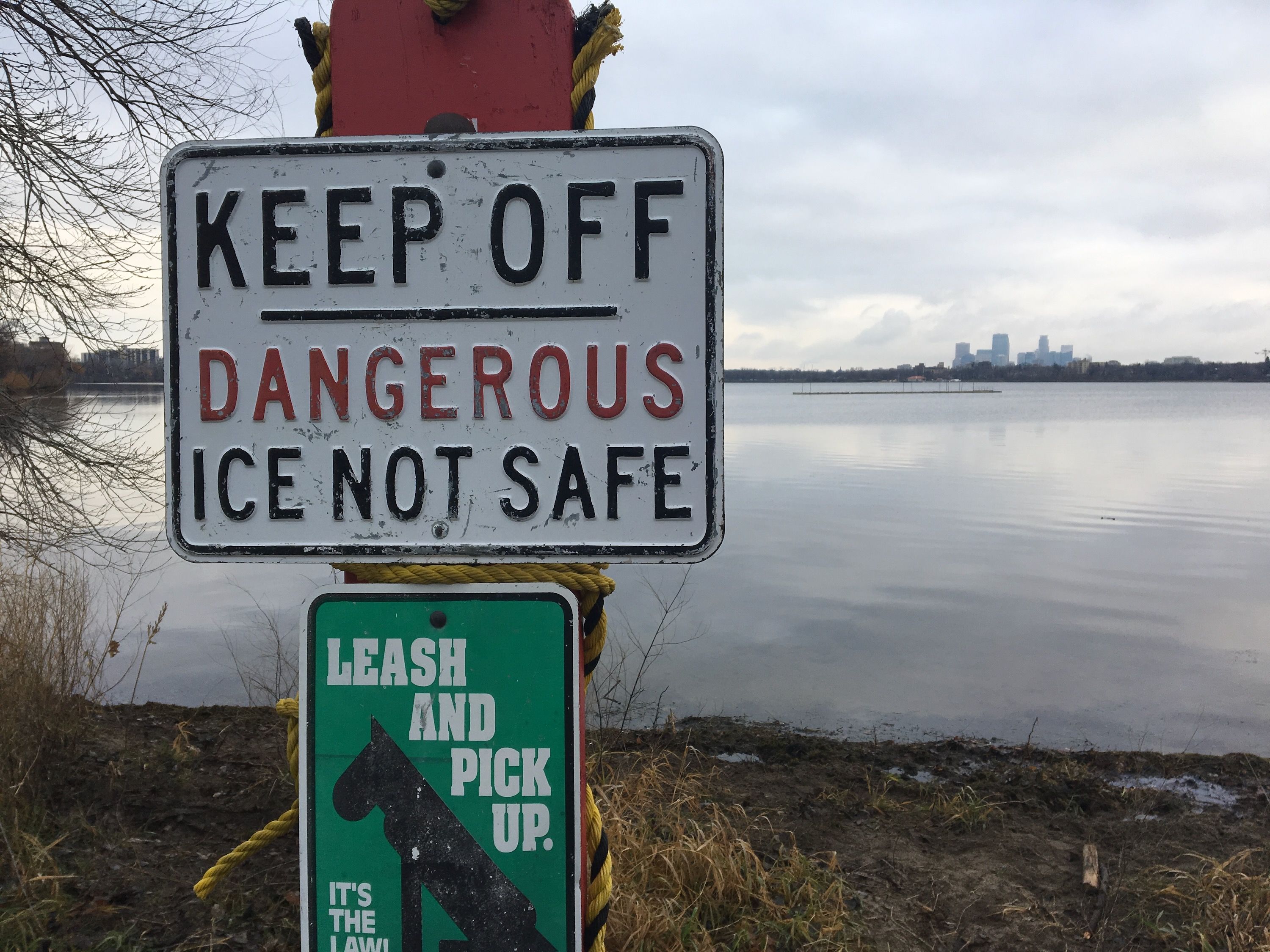 Weathered signpost by a lake with a large KEEP OFF and DANGEROUS ICE NOT SAFE sign, a green leash pickup panel; bare trees, calm water, and a distant city skyline.