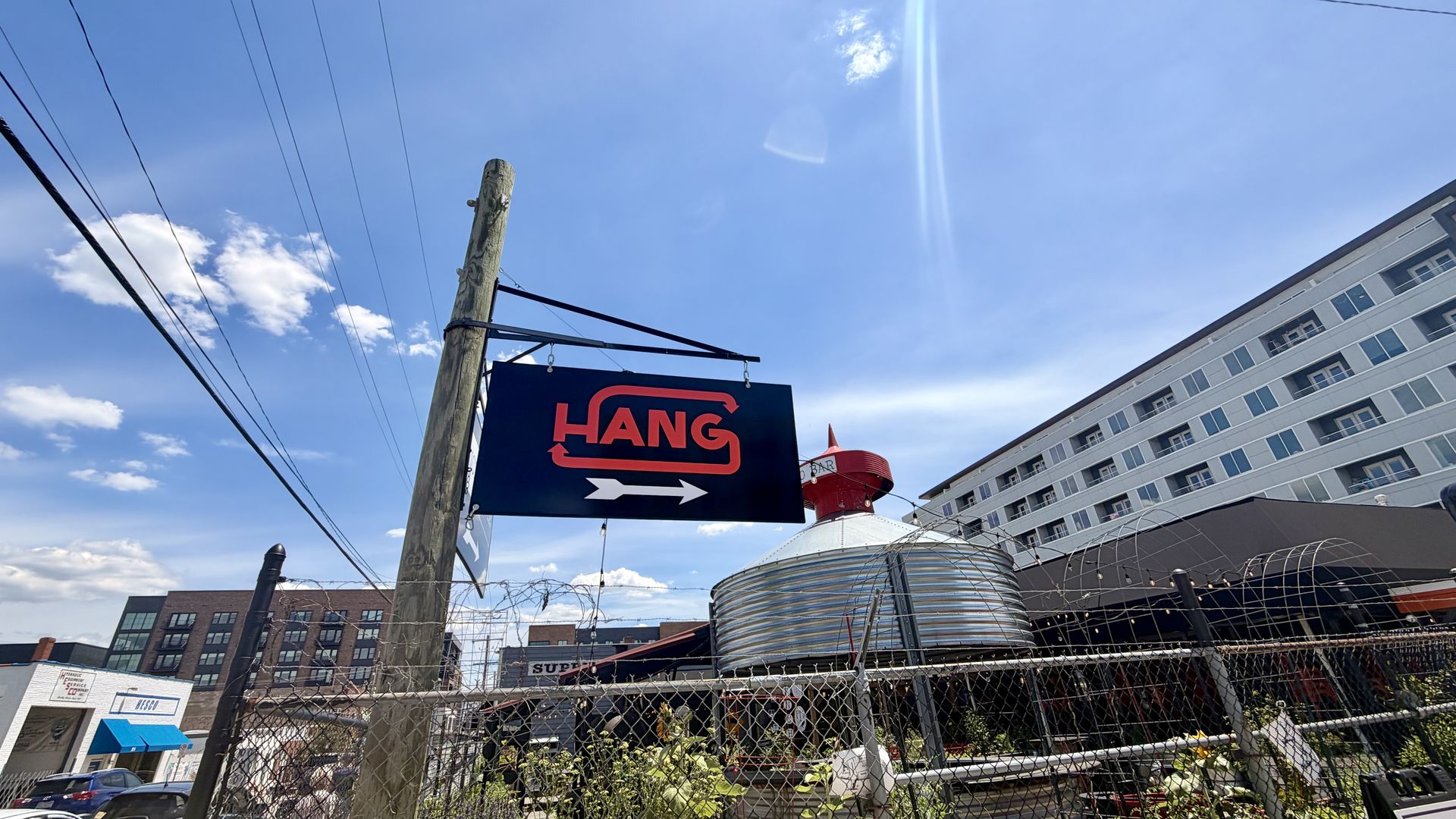 Blue sky with a wooden utility pole and a dark blue sign reading "HANG" in red with arrows. Overhead power lines, a gray apartment building on the right, and a chain-link fence in front.