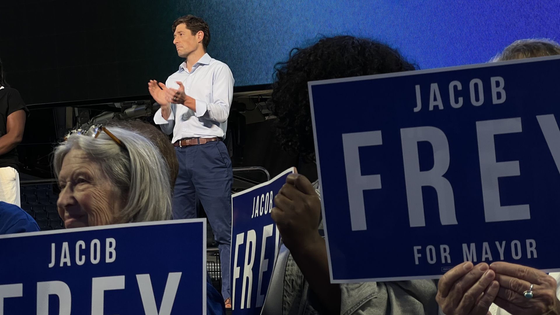 People holding blue signs that say "Jacob Frey for Mayor" at a political event, with a man in a white shirt and blue pants clapping on stage.