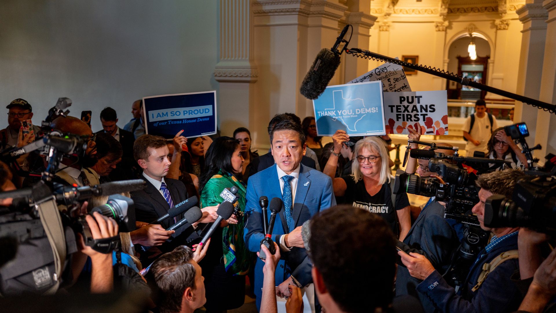 A man in a blue suit speaking to reporters inside a building, surrounded by people holding signs saying "I AM SO PROUD", "THANK YOU, DEMS!", and "PUT TEXANS FIRST" with cameras and microphones.