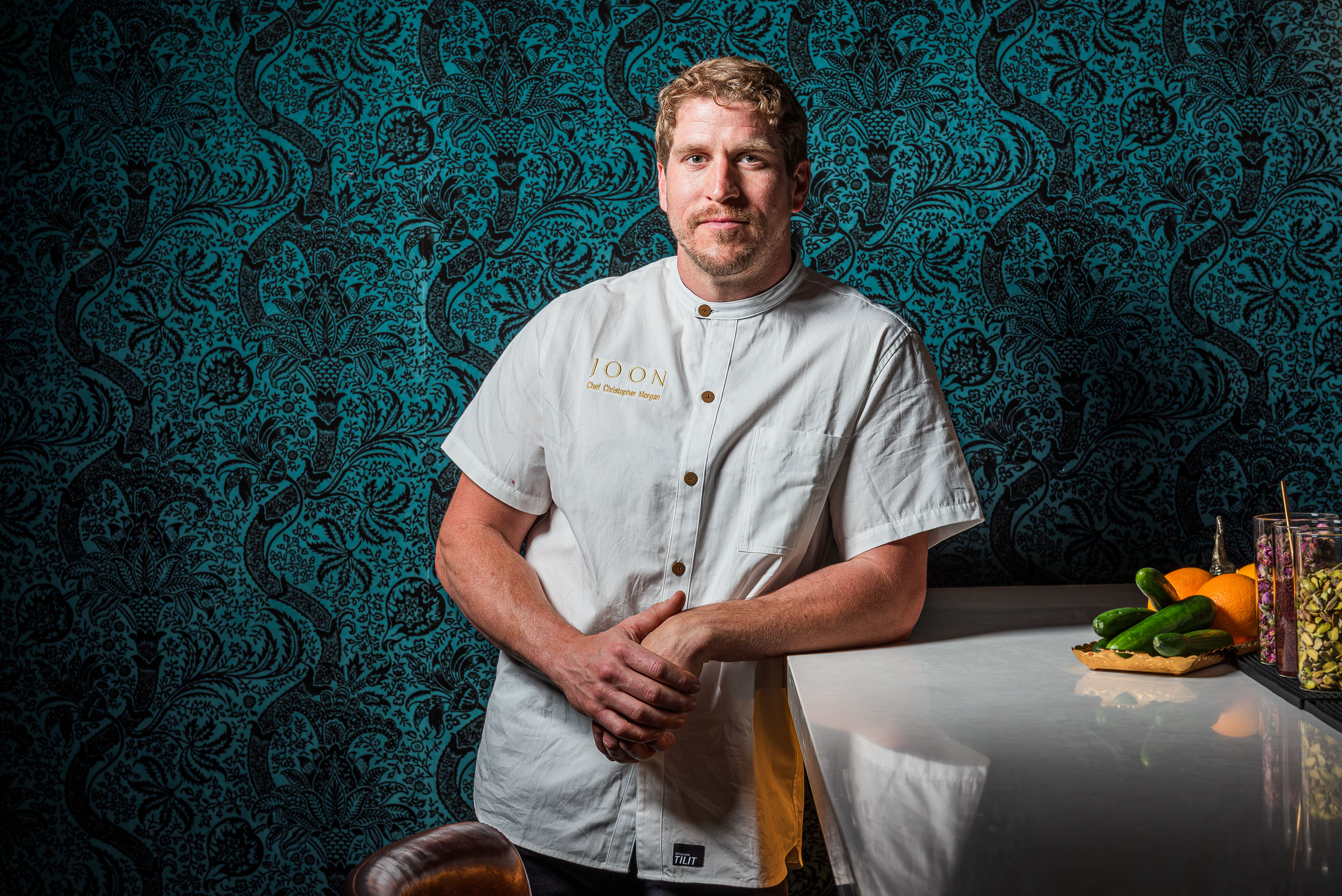 Chris Morgan in a white chef's jacket with text "JOON Chef Christopher Morgan" stands against a teal and black intricate patterned wall, leaning on a marble counter with cucumbers and oranges.