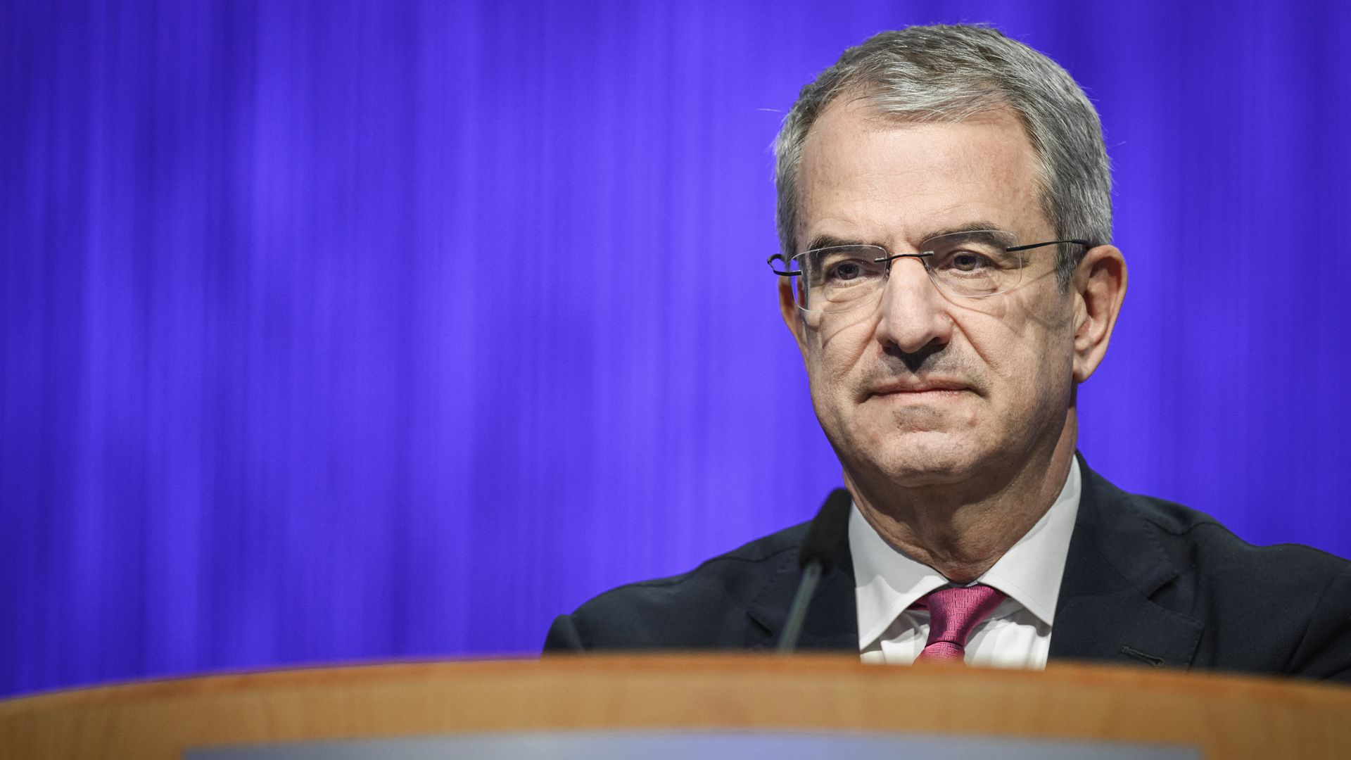 The gray-haired, bespectacled Nestlé CEO Laurent Freixe, wearing a black jacket, white shirt and red tie, stares ahead at a wooden podium, in front of a purple curtain.