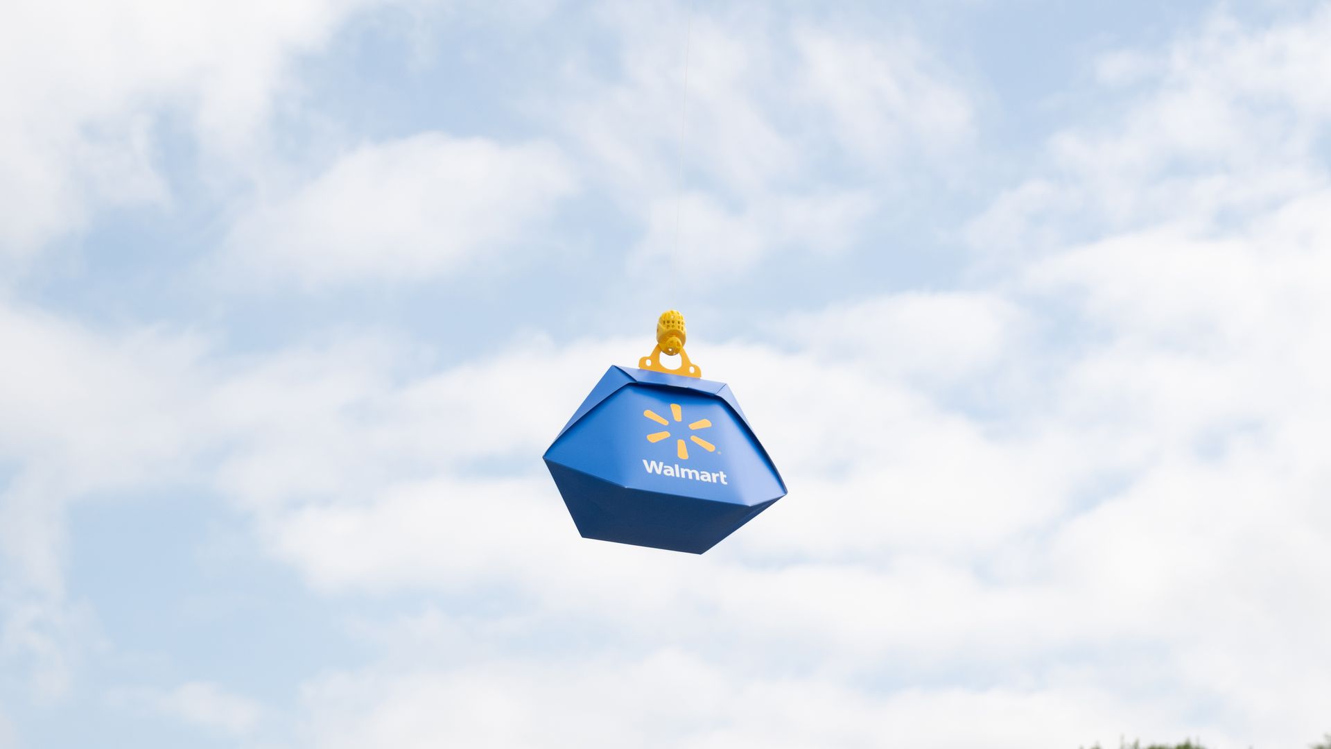 A Wing drone is lowering a blue Walmart branded package to the ground  against a partly cloudy sky with green tree tops visible at the bottom.