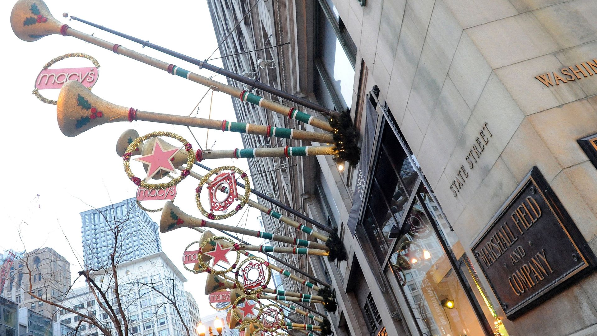 Festive holiday decorations with large trumpets and red stars hanging outside a building on State Street near Marshall Field and Company in a city with tall buildings.