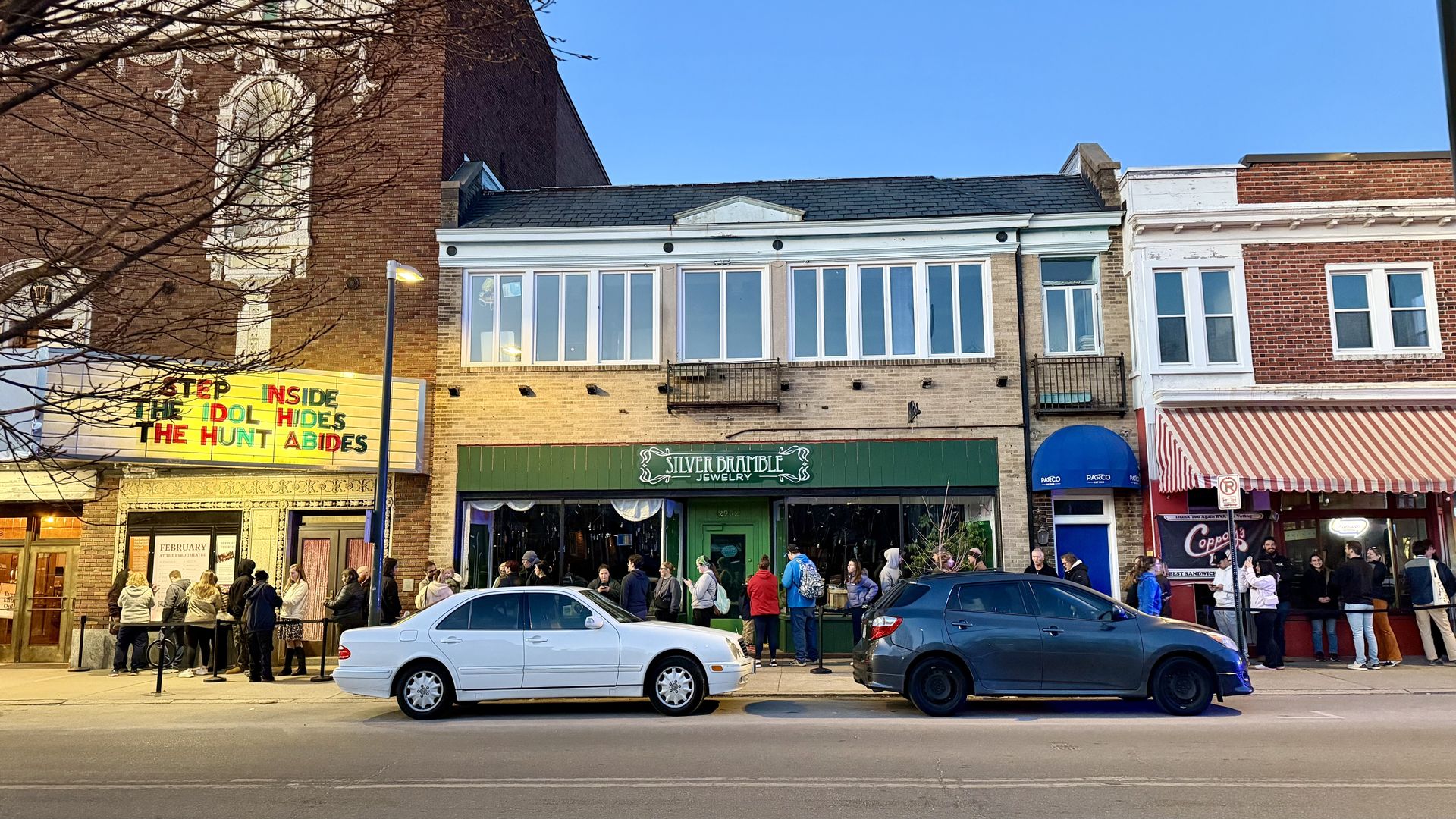 People line up outside a street with brick buildings at dusk, including Silver Bramble Jewelry with green awning and a theater with a colorful marquee saying, "Step inside the idol hides the hunt abides."
