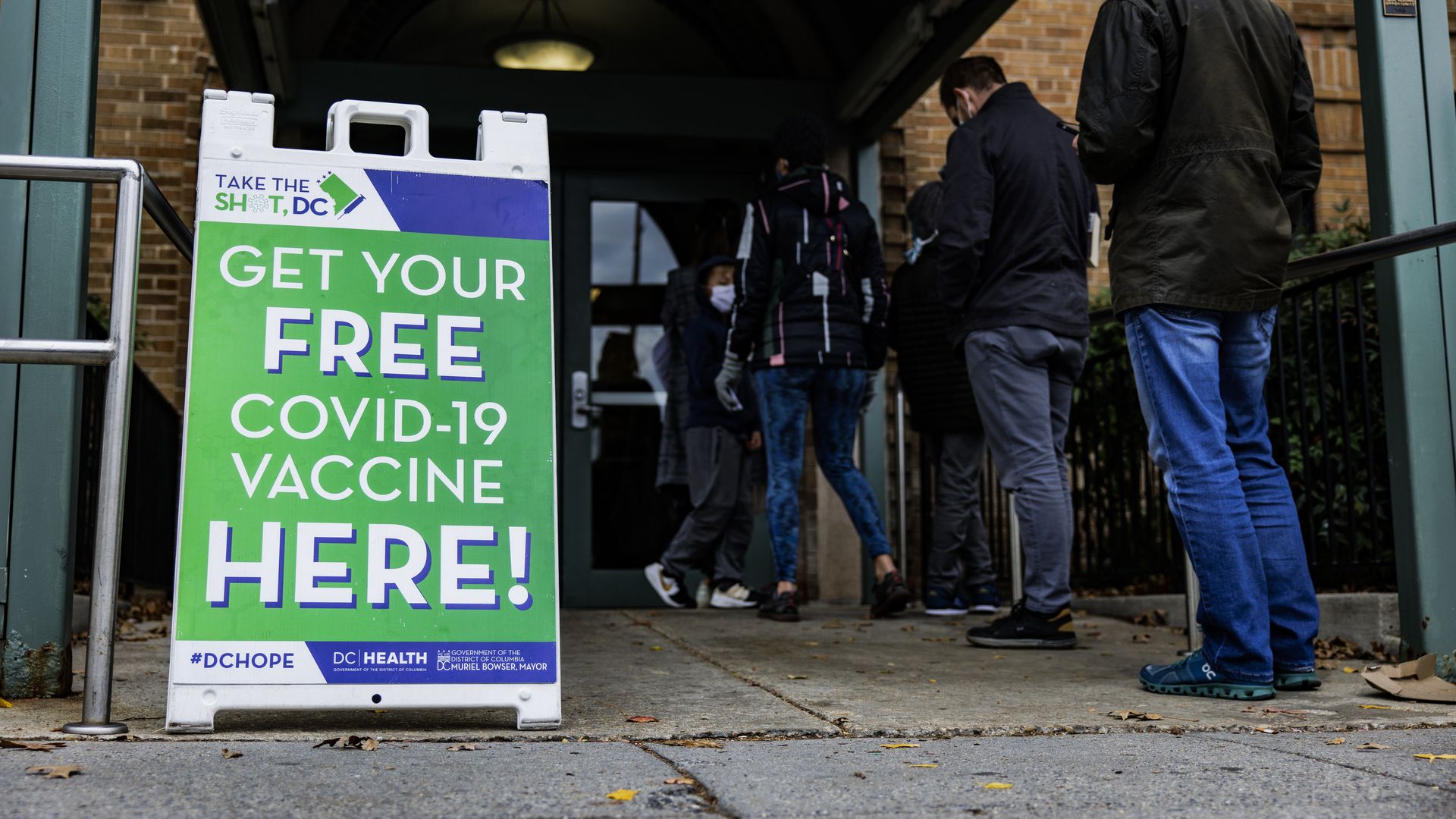 A line of people wait outside a building. A green sign is to the left of the line and says "get your free COVID-19 vaccine here!"