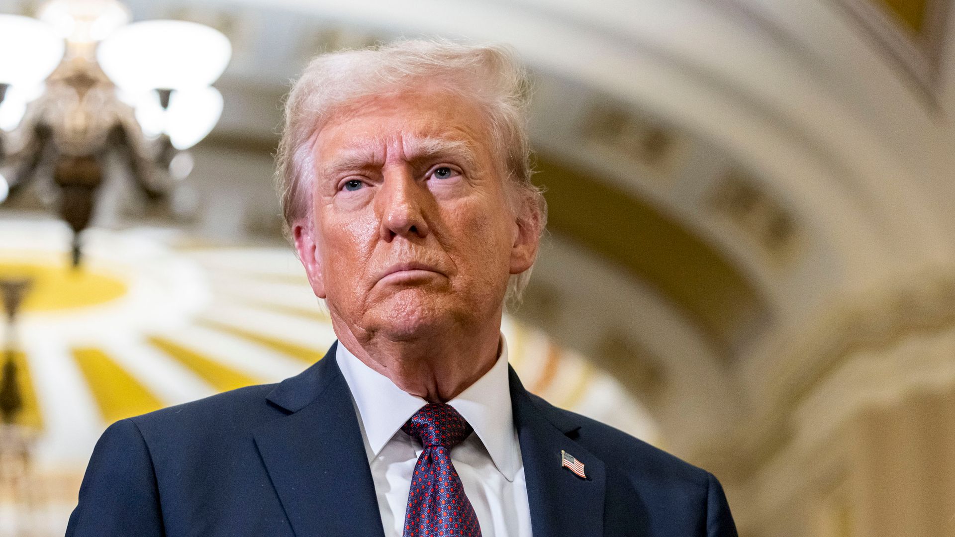 President-elect Donald Trump, wearing a blue suit and standing below a yellow and white ceiling.
