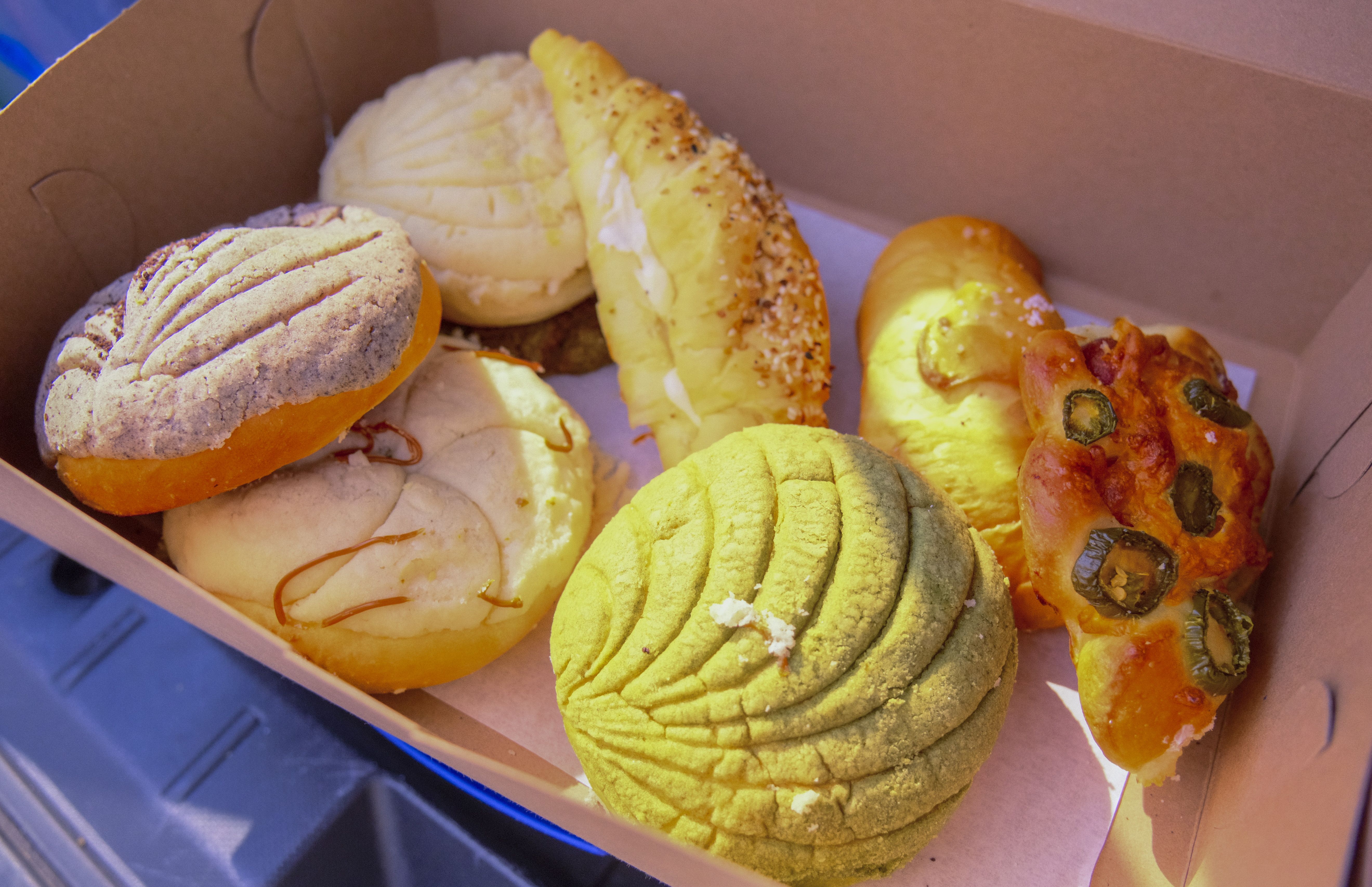 Box with assorted Mexican pastries including conchas in cream, brown, and green colors, a sesame-covered bolillo, and a jalapeño-topped savory bread slice in natural light.