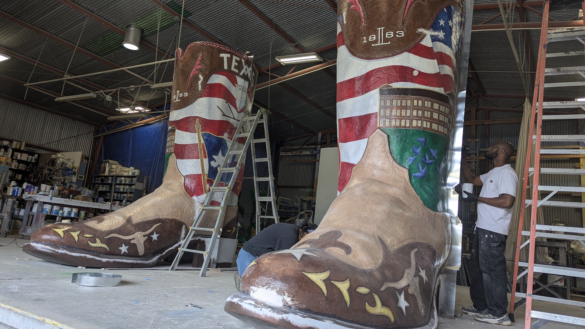 Two giant painted cowboy boots featuring Texas designs inside a large industrial workshop with two people working near them and ladders nearby.