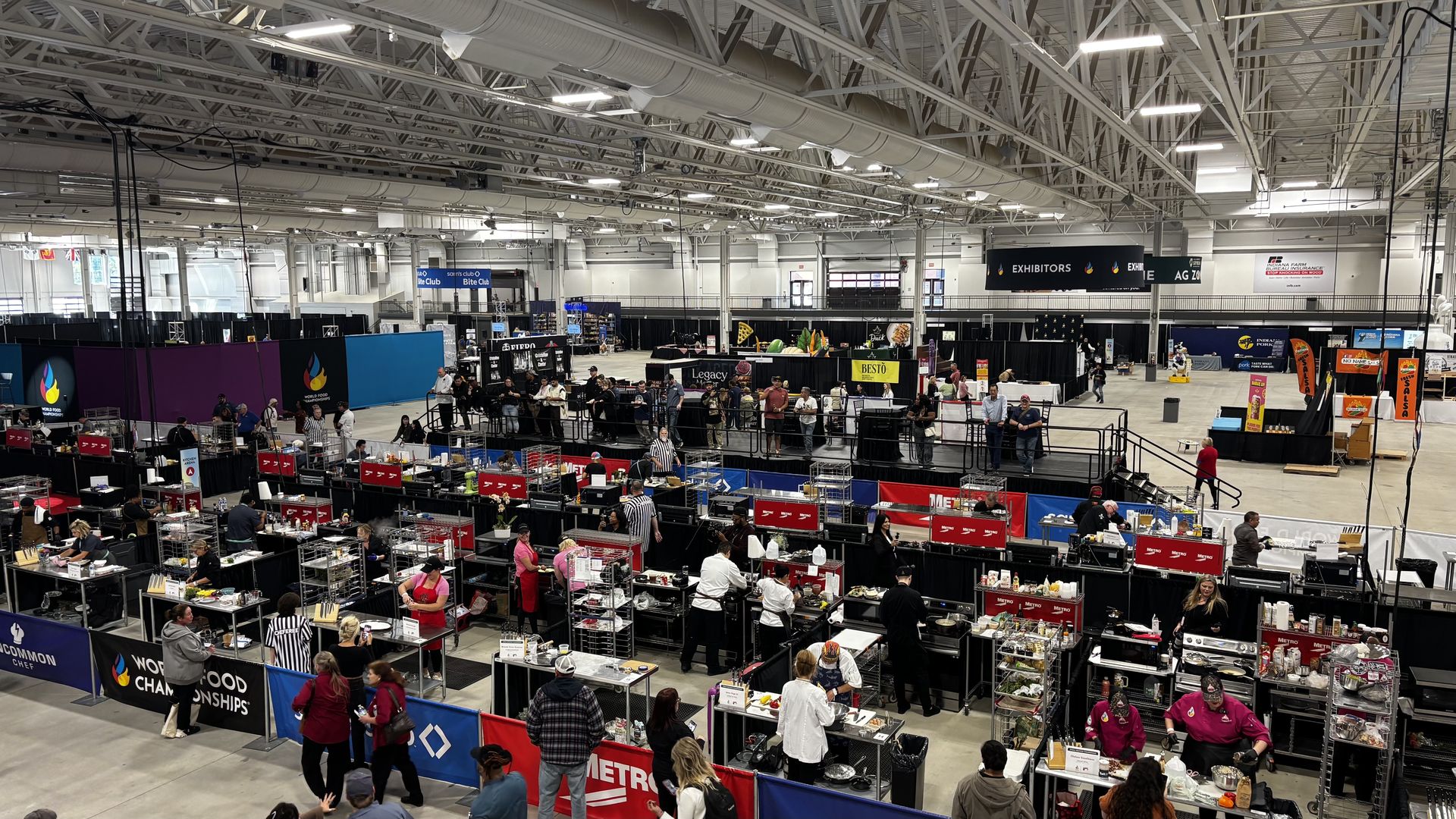 Inside a large convention hall, rows of vendor booths and display tables line wide aisles. People browse cooking gear and food displays beneath bright ceiling lights and exposed ductwork.