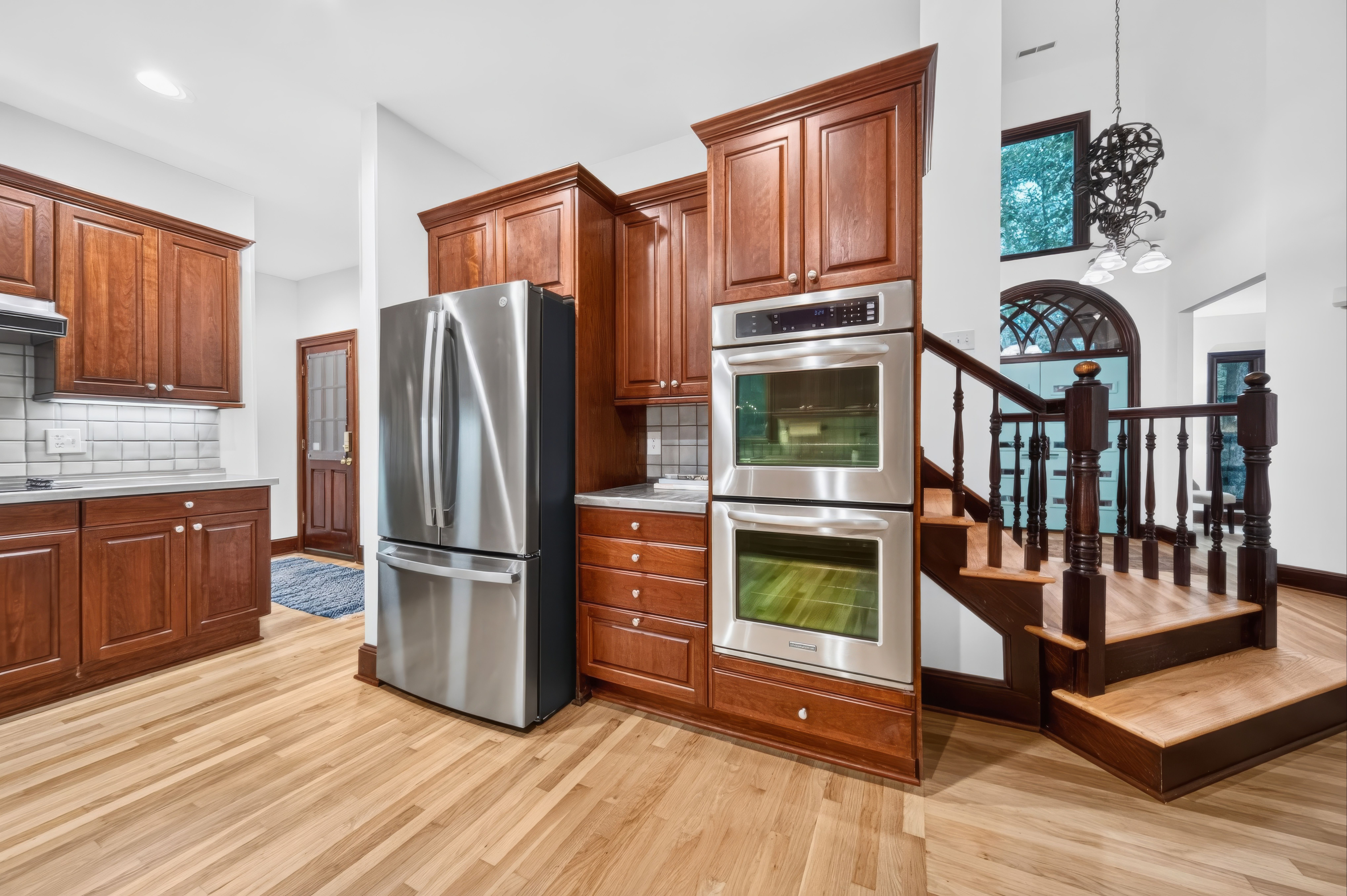 Kitchen with dark wood cabinets, light hardwood floors, and a stainless steel refrigerator with double ovens. White walls, dark wood staircase railing, an arched window, and pendant lights.