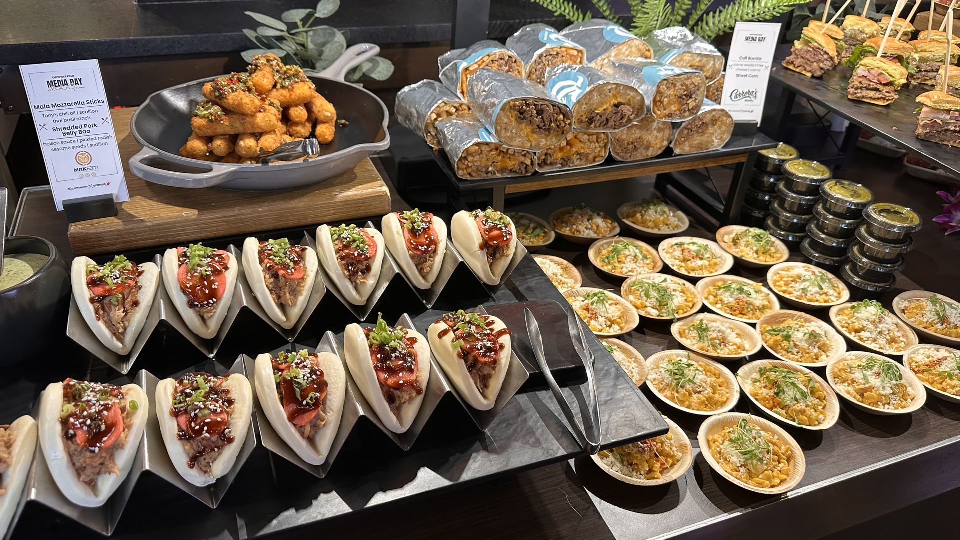 Catering display with a skillet of crispy croquettes topped with sauce, foil-wrapped burritos stacked on a tray, row of mini buns with meat and greens, and bowls of colorful salads and grains.