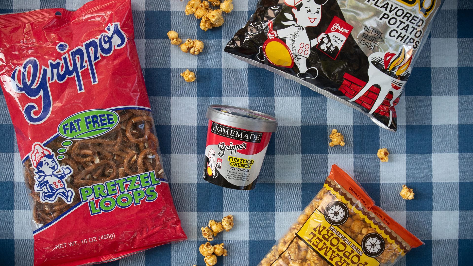 Top-down view of snacks on a blue checkered table: a red bag of pretzel loops on the left, a black-and-white chip bag on the right, scattered popcorn, and a small can labeled Homemade in the center.