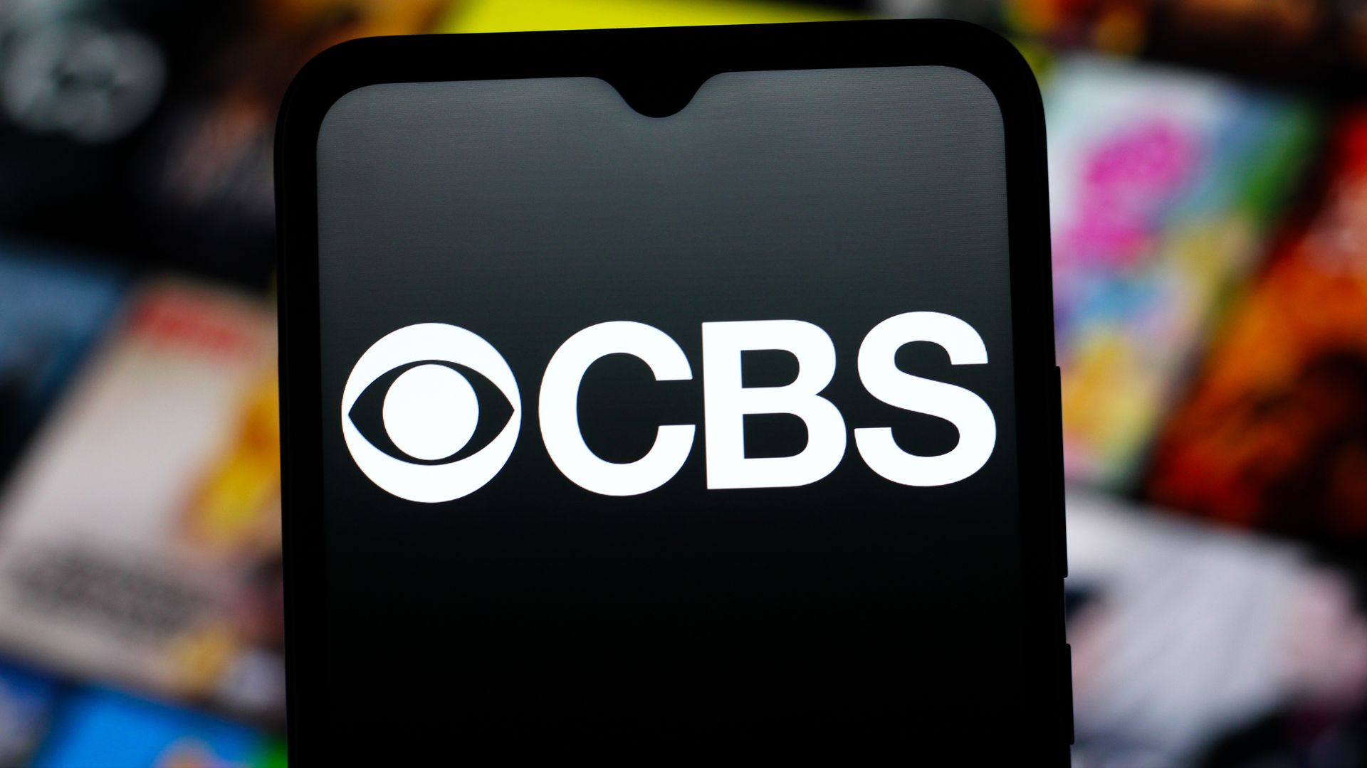 A smartphone screen displaying the white CBS logo on a black background with colorful blurred objects in the background.