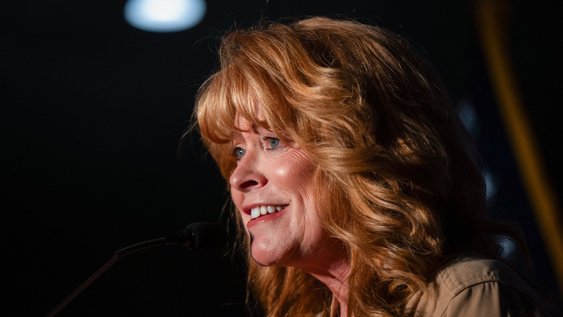 Close-up of a smiling woman with light brown hair speaking into a microphone on a dark stage. Warm lighting highlights her face and hair, with a blurred blue light in the background.