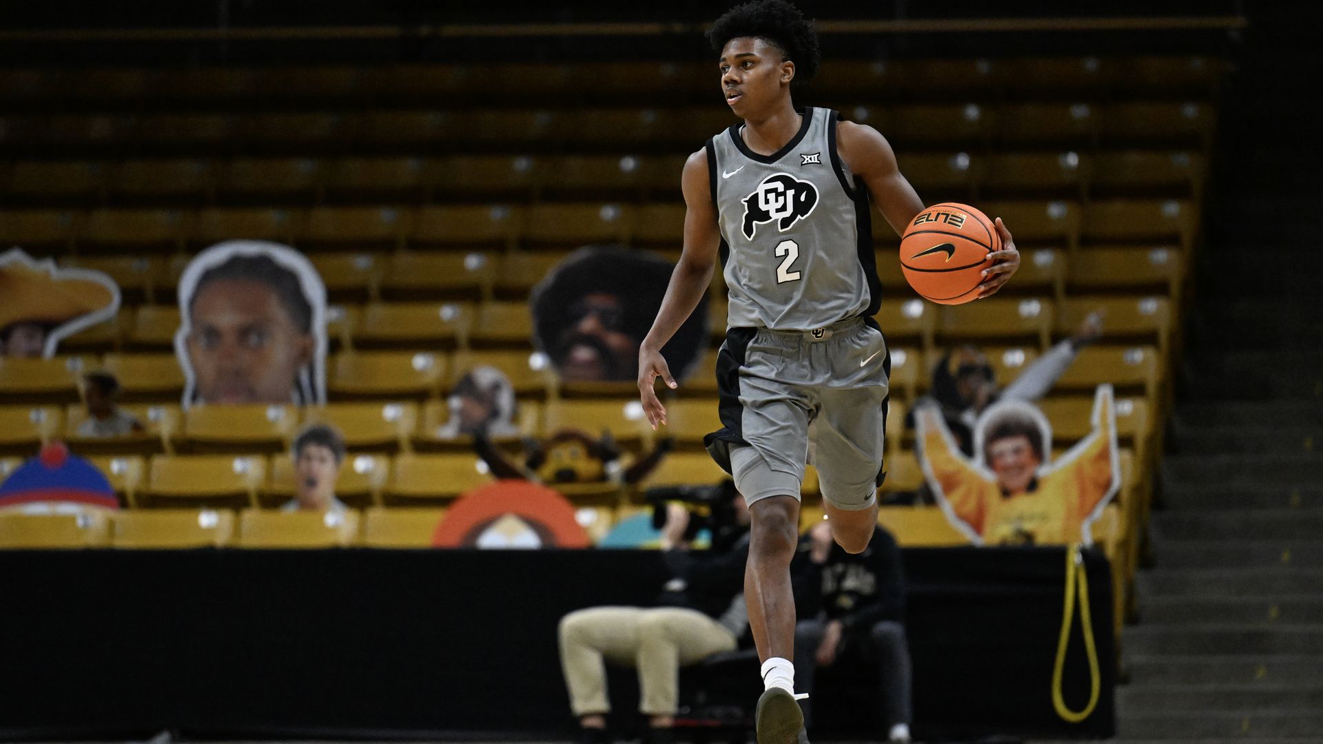 Basketball player in gray Colorado uniform dribbling an orange basketball on court with empty yellow seats and large face cutouts behind him.
