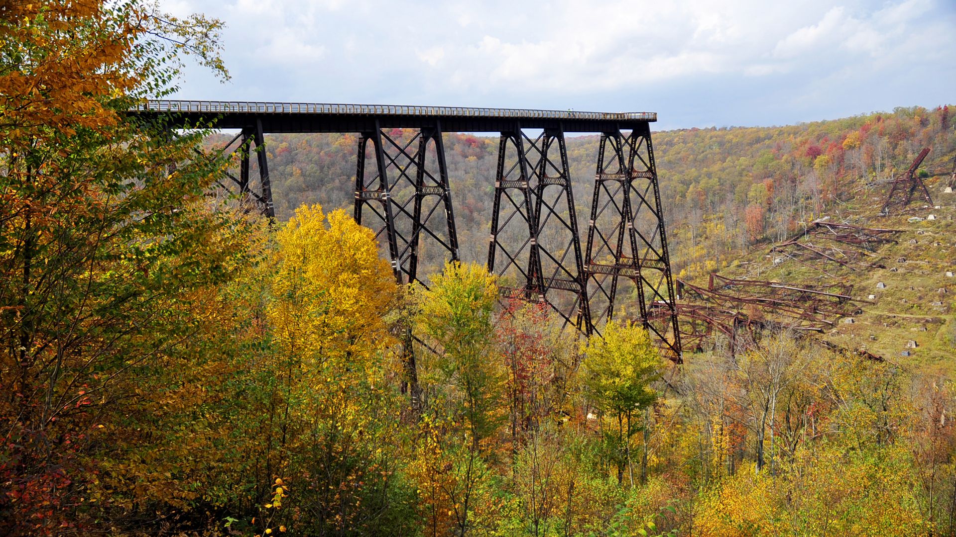 Tall rusty railway trestle bridge over a forested valley with colorful autumn foliage, partly collapsed structure to the right under a cloudy sky.