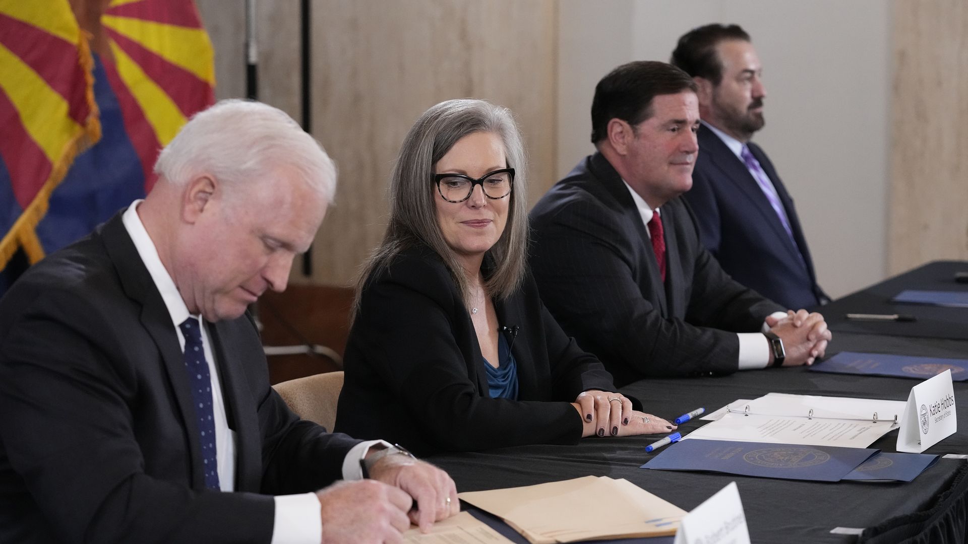 Robert Brutinel, Katie Hobbs, Doug Ducey and Mark Brnovich are seated side by side at a long table while Brutinel signs a document