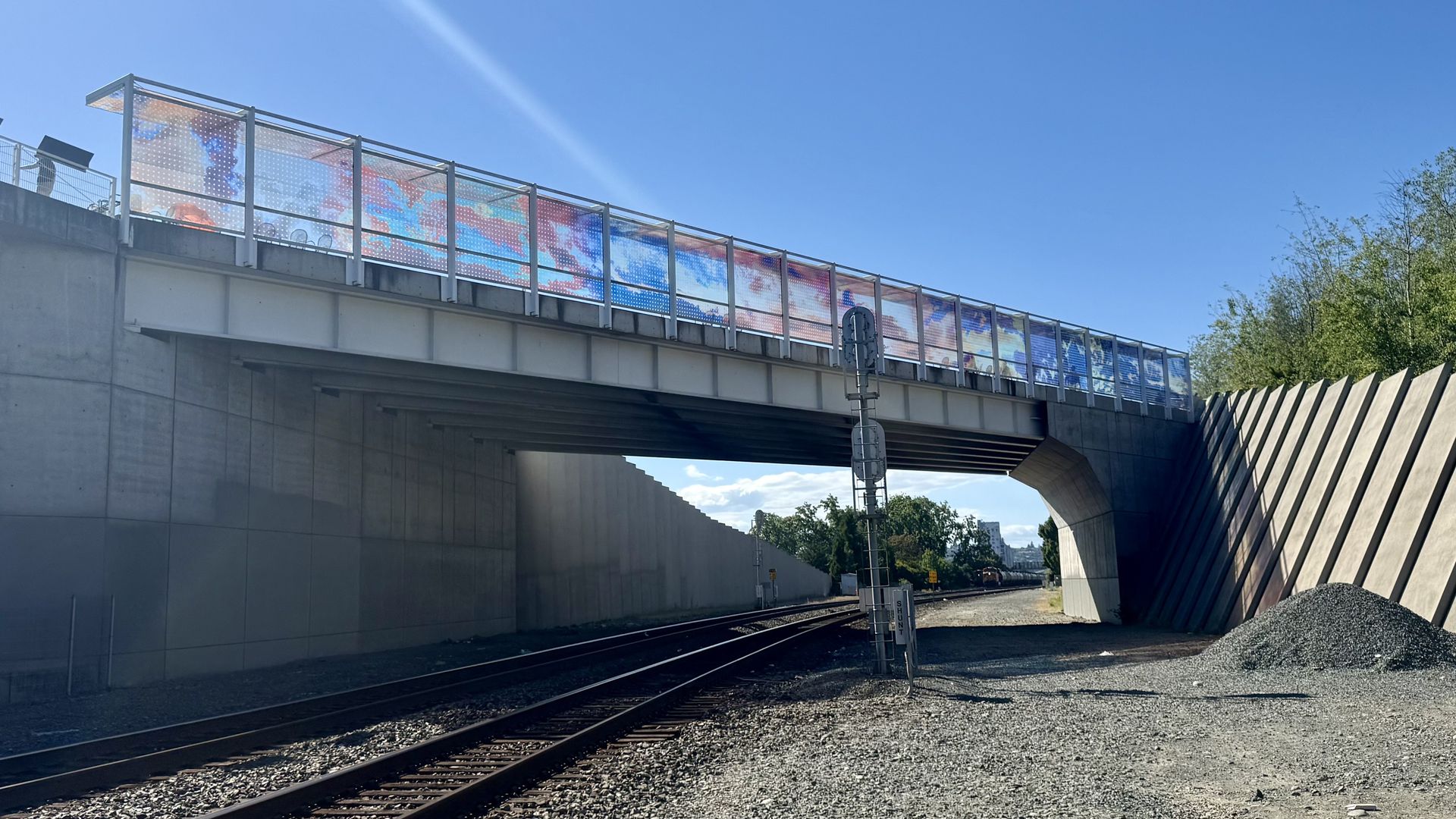Train tracks pass beneath a concrete overpass featuring colorful art panels along the railing under a clear blue sky.