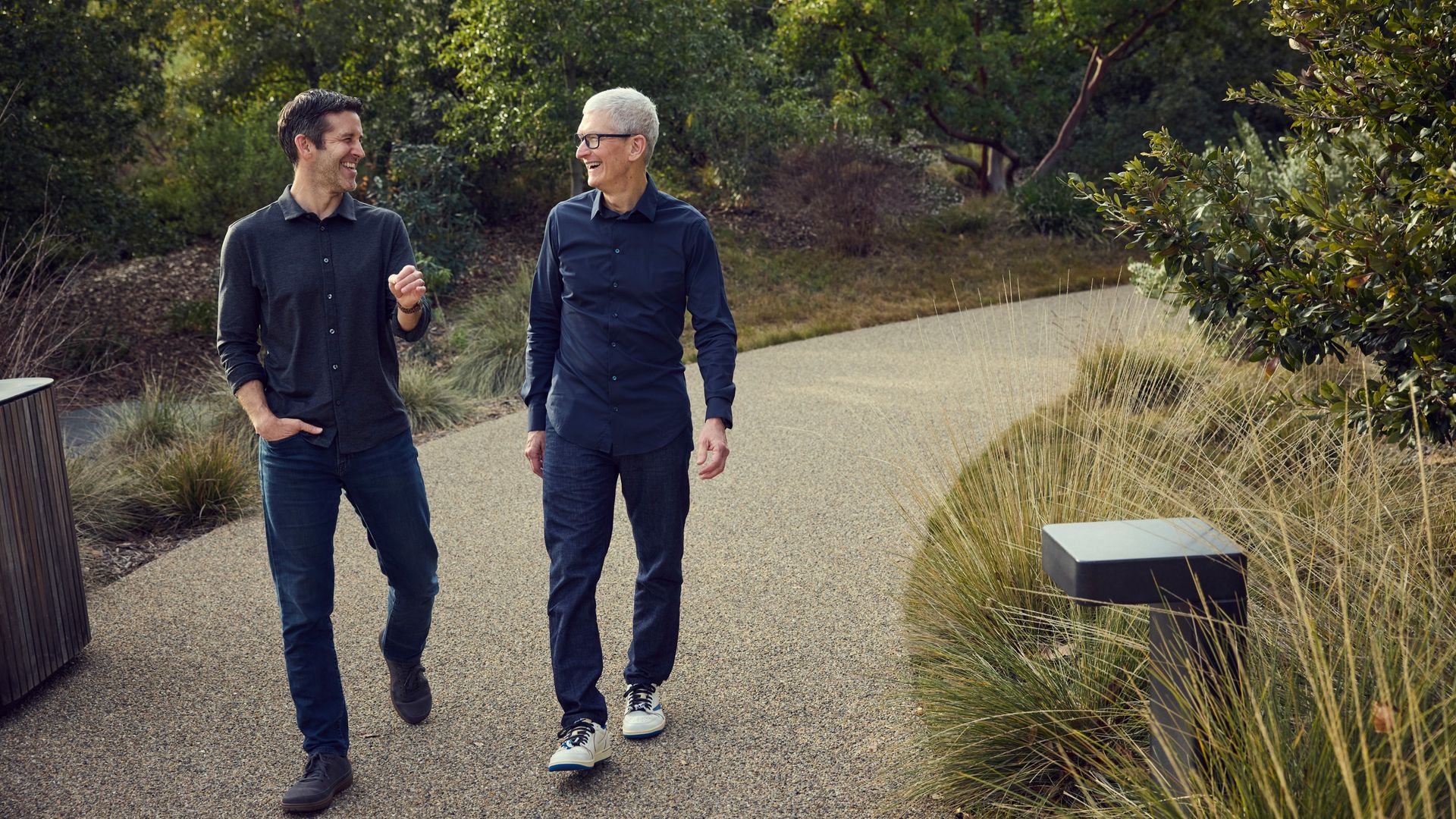 Two men walk and chat along a sunny park path, smiling. One in a dark gray shirt, one in a navy shirt with glasses, surrounded by green trees and ornamental grasses.