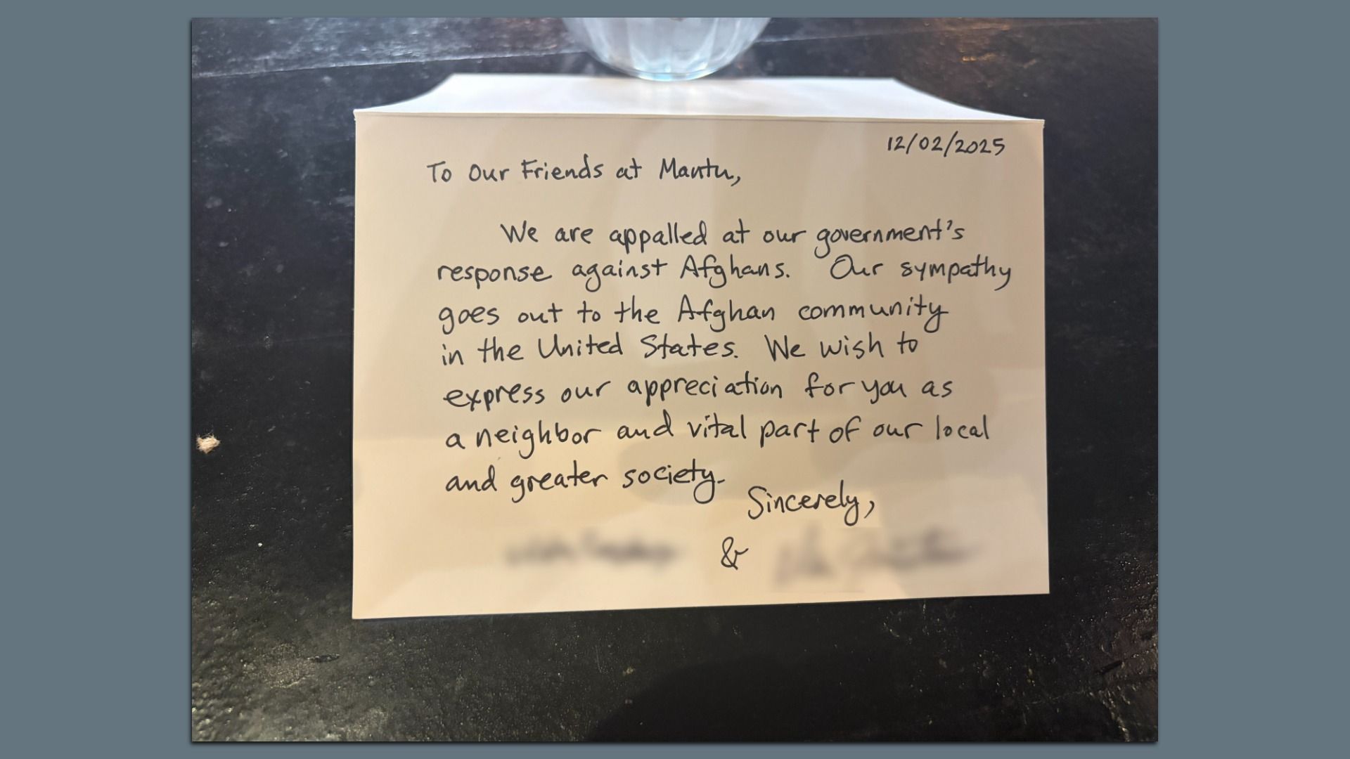 Handwritten letter dated 12/02/2025 expressing sympathy for Afghans and appreciation for a local Afghan community, addressing friends at Mantu, on a dark surface beneath a glass.