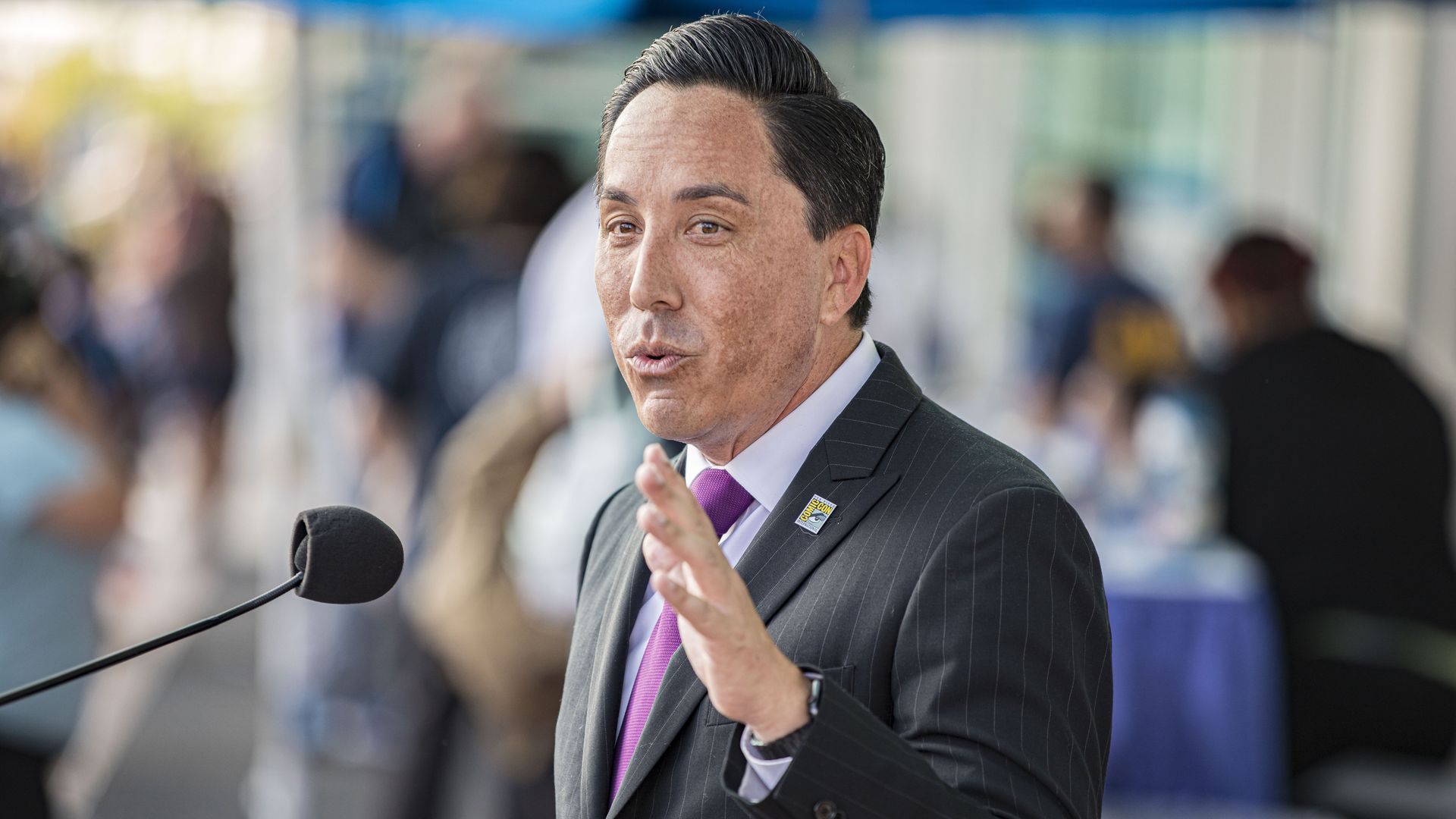 Mayor Todd Gloria wearing a pinstriped suit and purple tie talking into a microphone