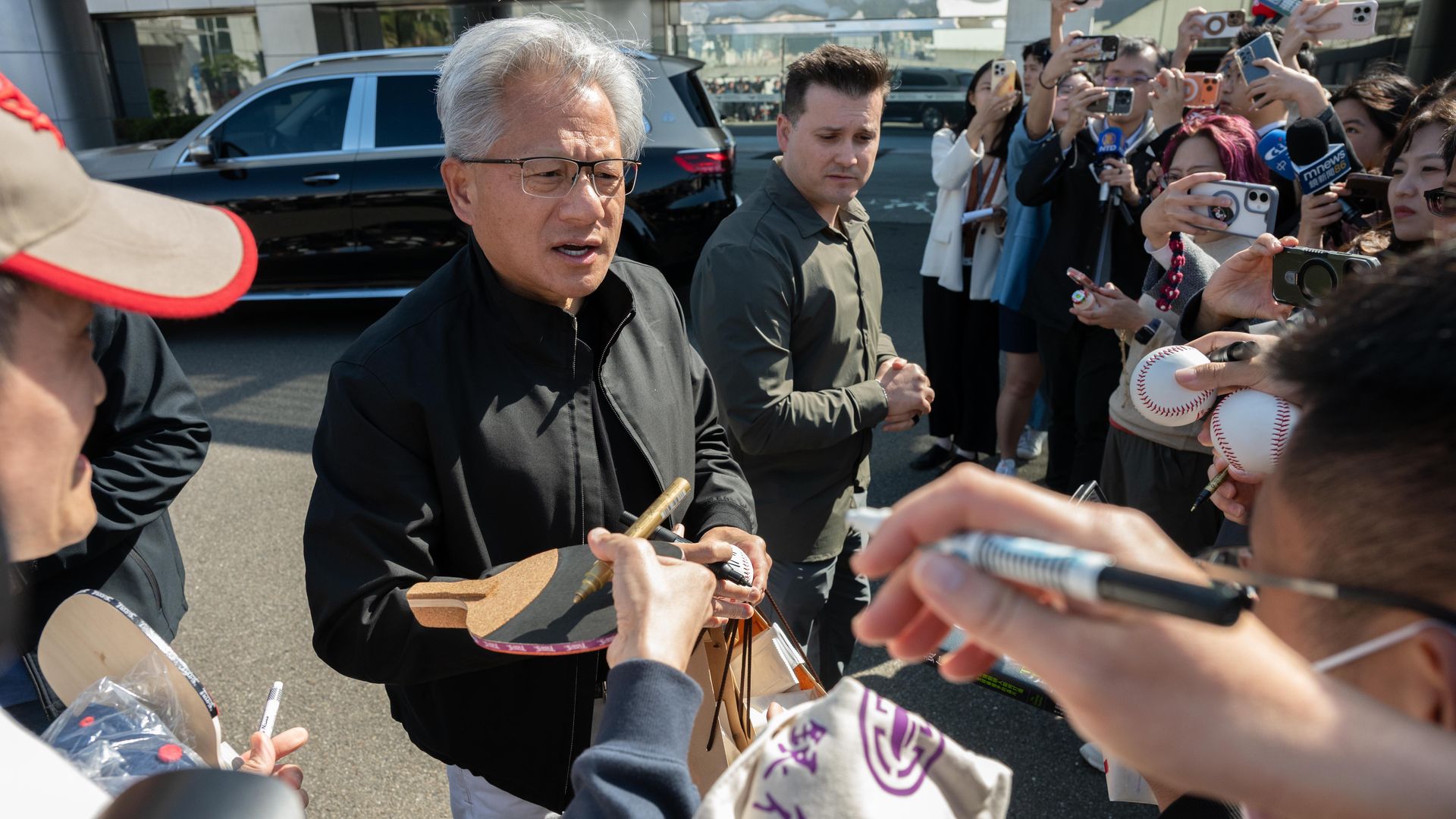 A gray-haired person in a black jacket signs autographs while surrounded by a crowd holding cameras and microphones