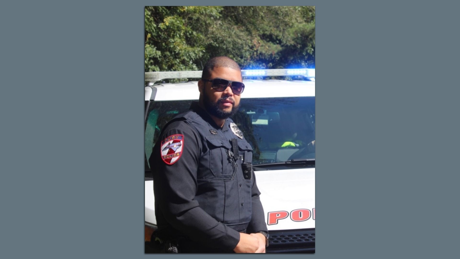 Police officer in black uniform and sunglasses stands with arms crossed in front of a white police vehicle with blue lights on top, trees in the background.