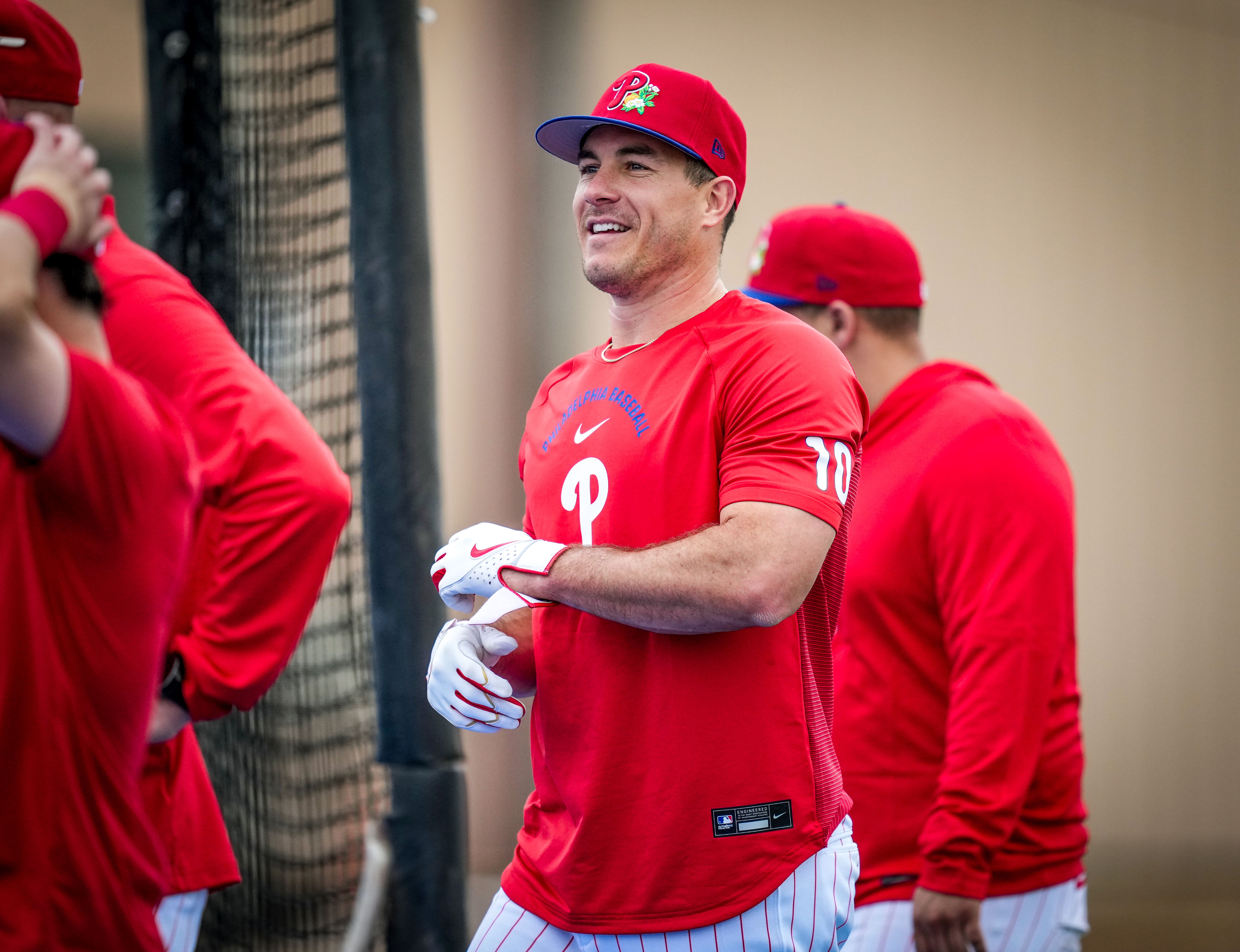 Smiling baseball player in red Philadelphia Phillies shirt and cap stands near batting cage, wearing white gloves, with teammates in red behind him.