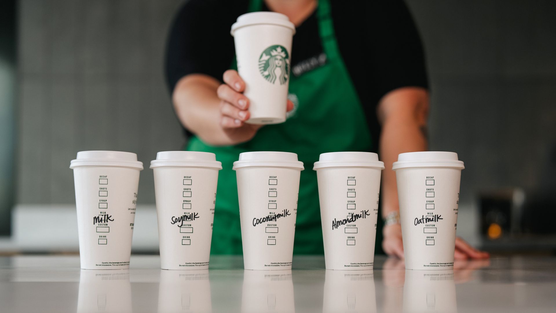 Starbucks cups lined up with the type of milk marked on the cups. A barista with green apron holds a cup with the green Starbucks logo above the row of five cups