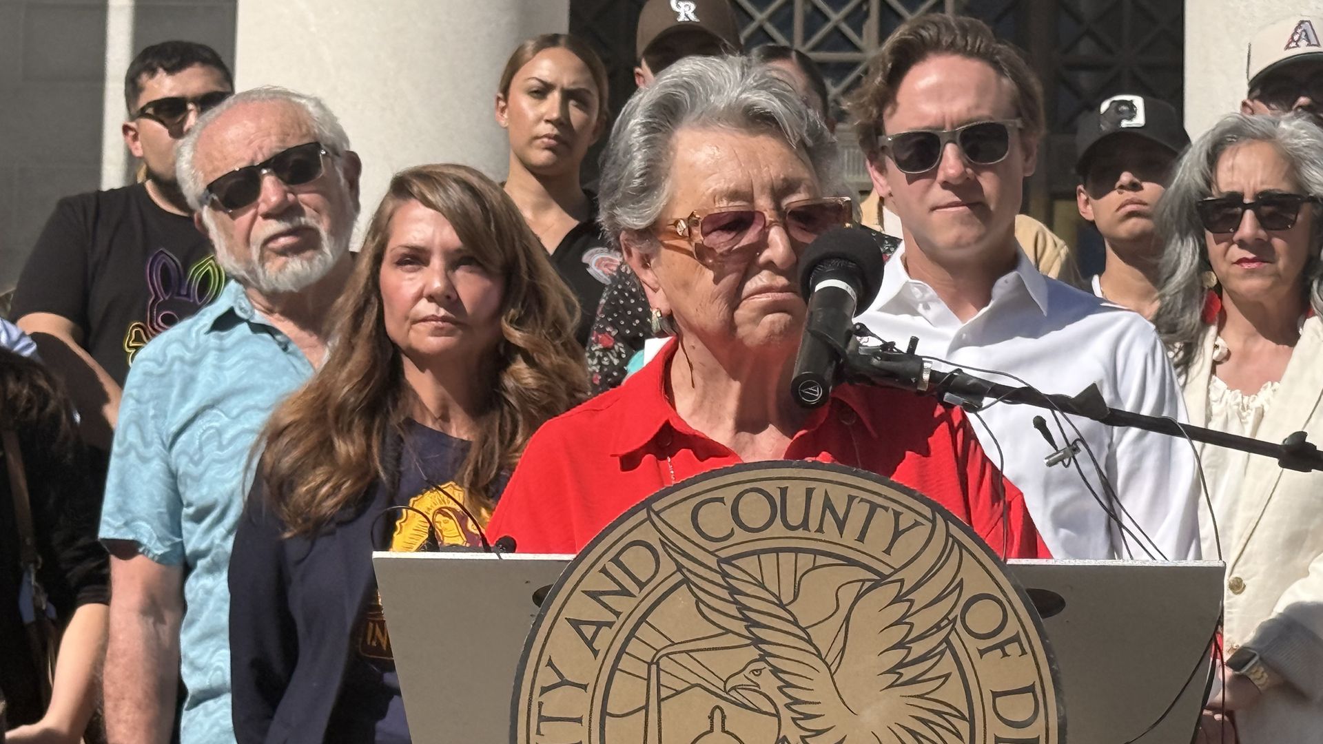 Older woman in a red shirt speaks into a microphone at a podium with a large circular seal; a diverse crowd stands behind her in daylight, many wearing sunglasses.