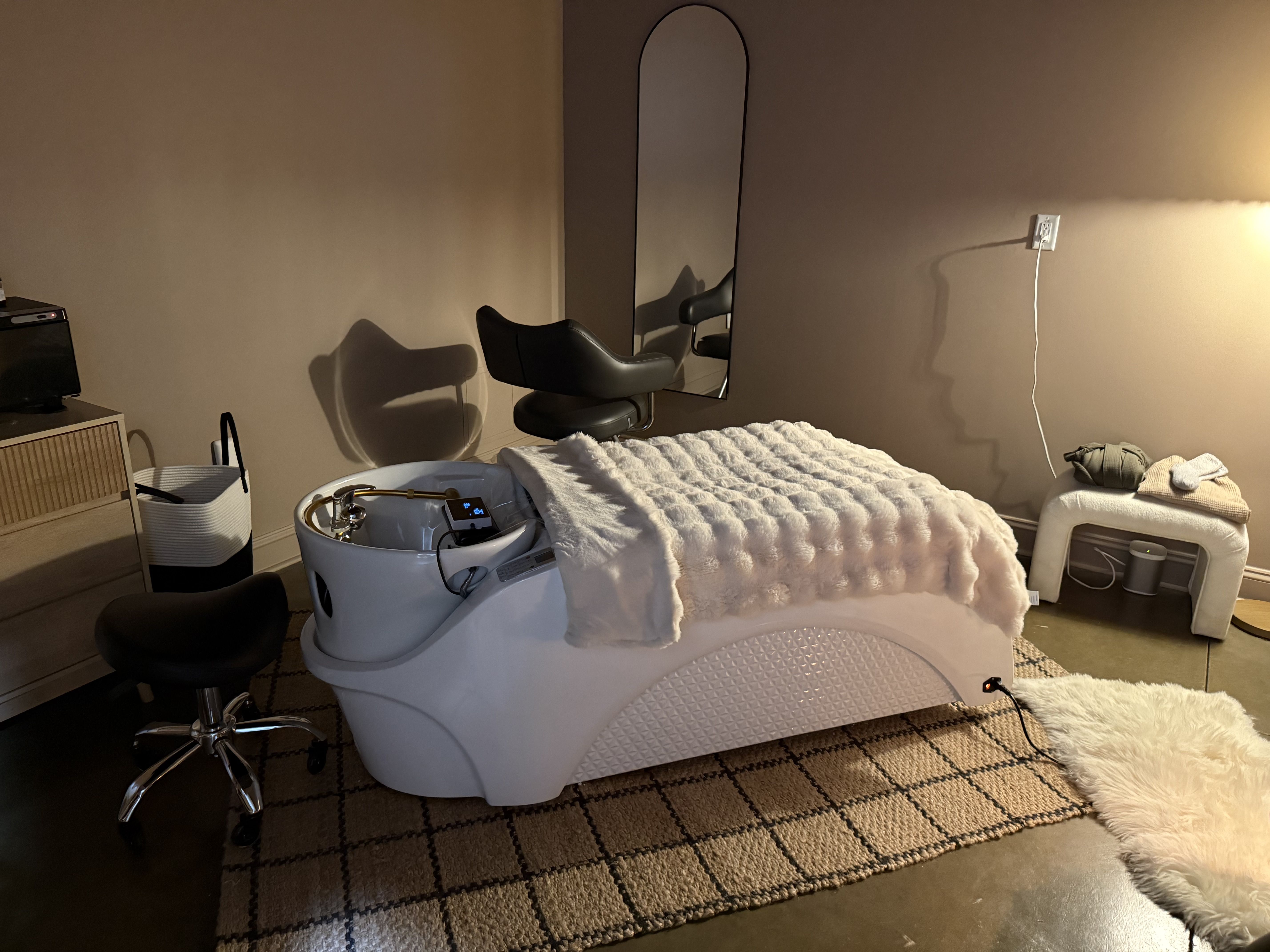 Dimly lit bedroom spa with a white pedicure chair and foot basin on a fluffy white blanket, a tall arched mirror on beige walls, a black rolling stool, and a power cord by the outlet.