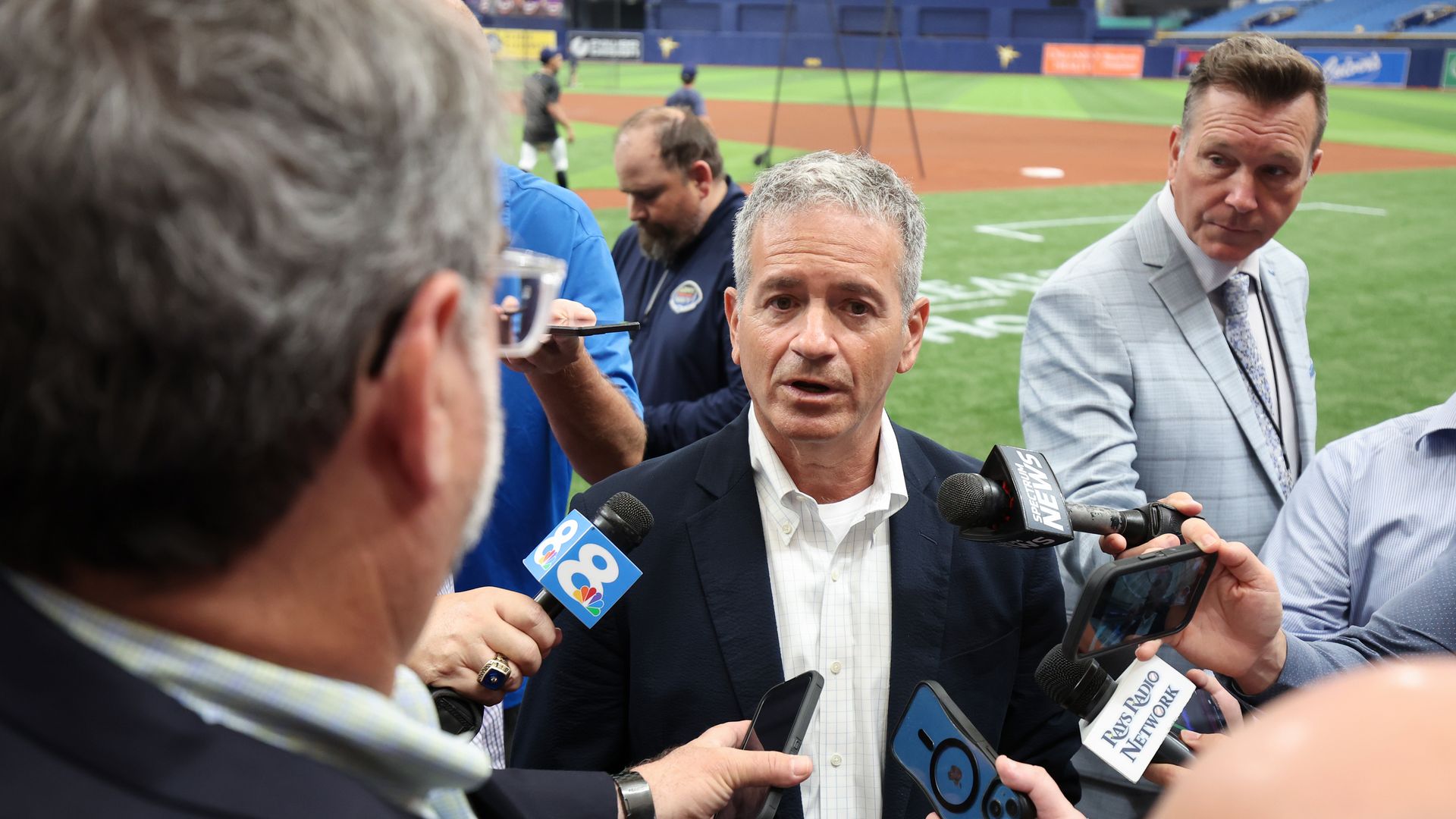 A man with gray hair and a navy suit talks to several reporters holding out microphones. In the background is a baseball field.