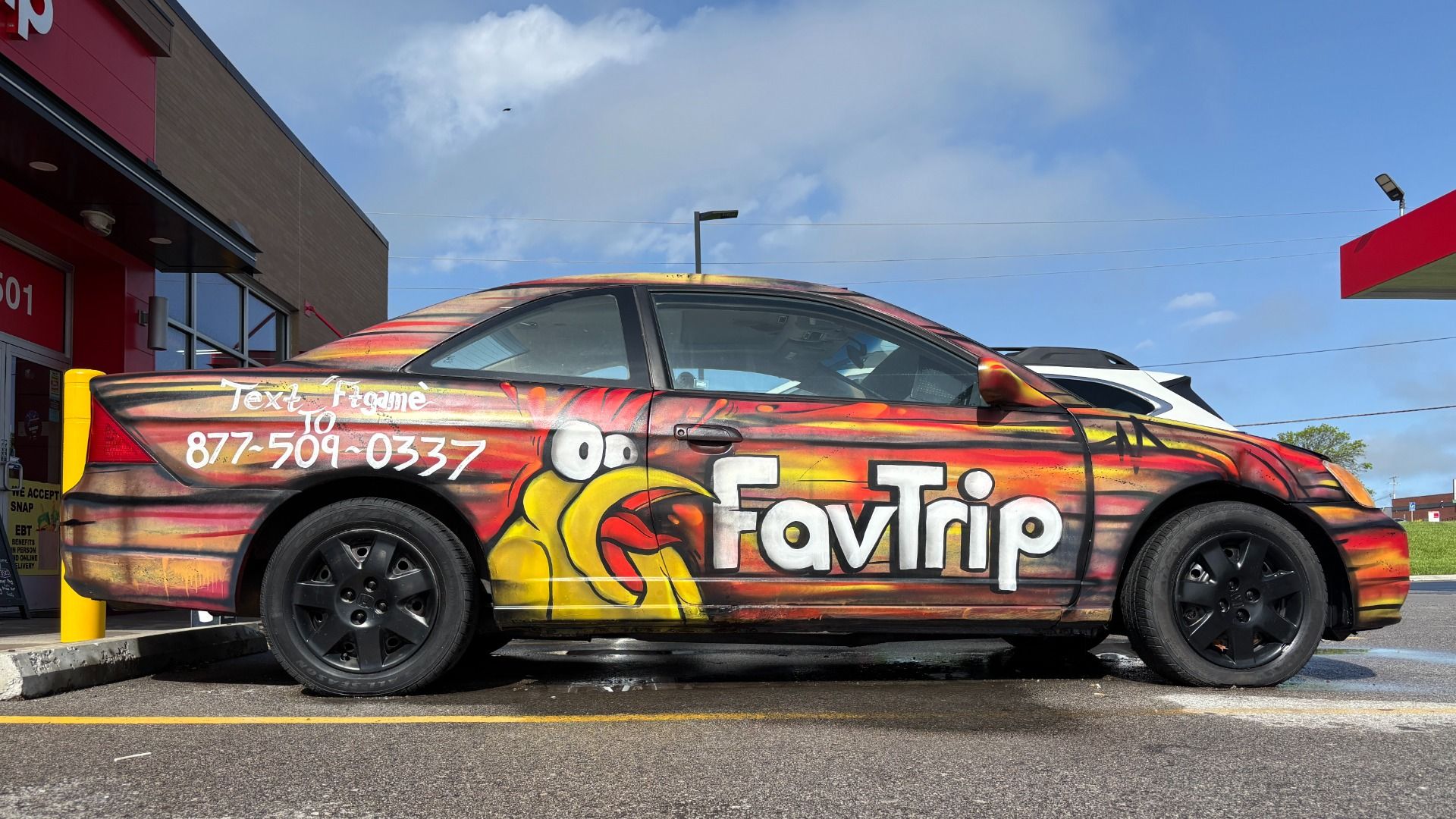 Colorful flame-wrapped car at a gas station with red, orange, and yellow flames, a cartoon yellow beak bird, "FavTrip" and the phone number 877-509-0337 on the side; blue sky.