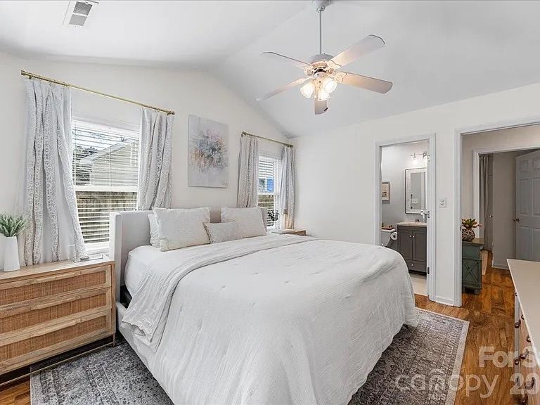 Bright white bedroom with king-sized bed, white bedding, wooden nightstand, two windows with light gray curtains, ceiling fan, and a door leading to a bathroom and adjoining room.