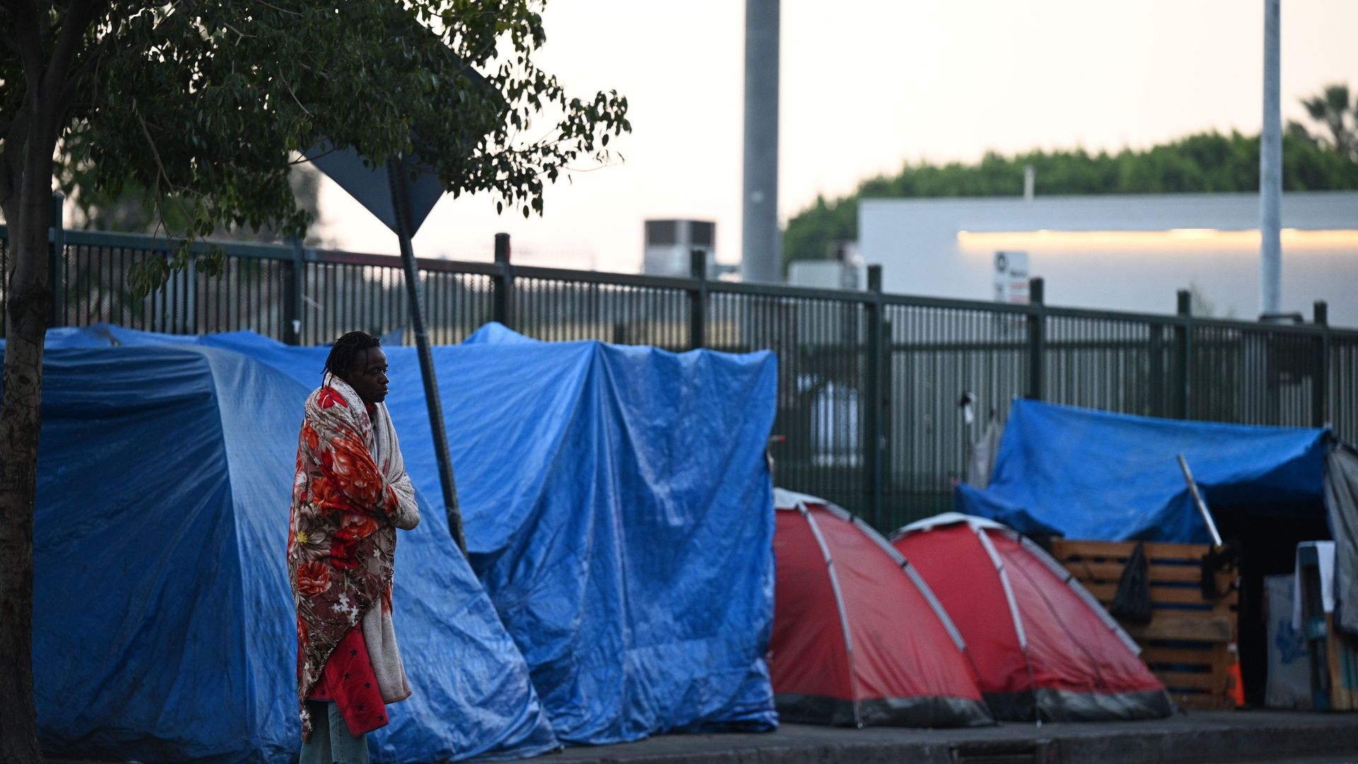A man wrapped in a blanket stands outside blue tarp tents 
