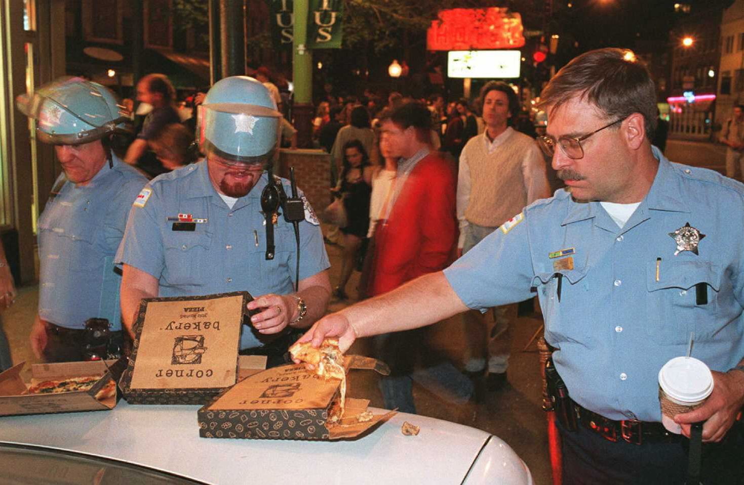 Night street scene with three light-blue police officers, two wearing helmets, around a car hood; one officer reaches for pizza from an open box while a crowd watches in the background.