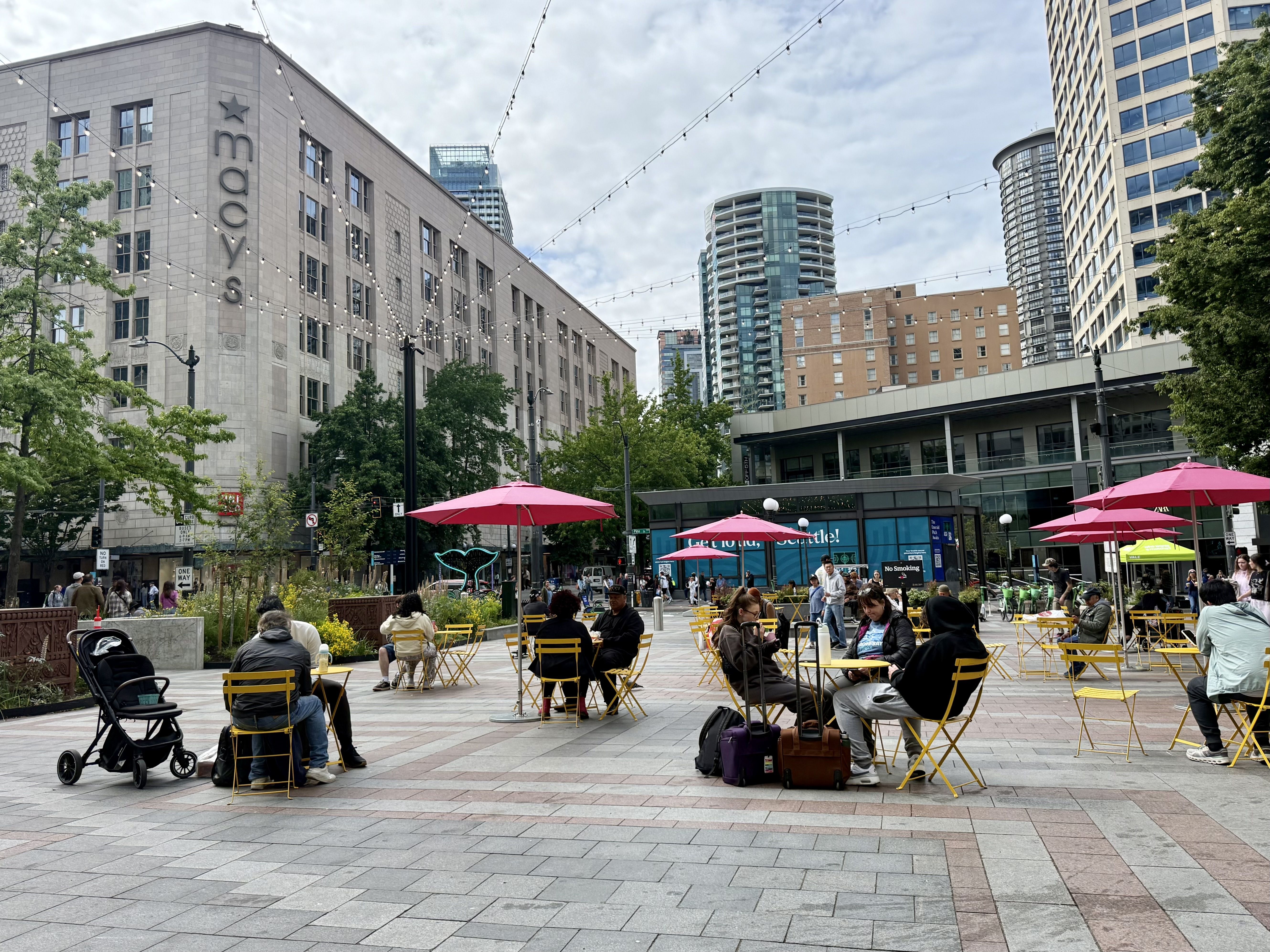Outdoor city square with string lights, umbrellas, cafe tables and yellow chairs. People sit and stroll near a stone Macy's building, with modern high-rises in the background under a cloudy sky.