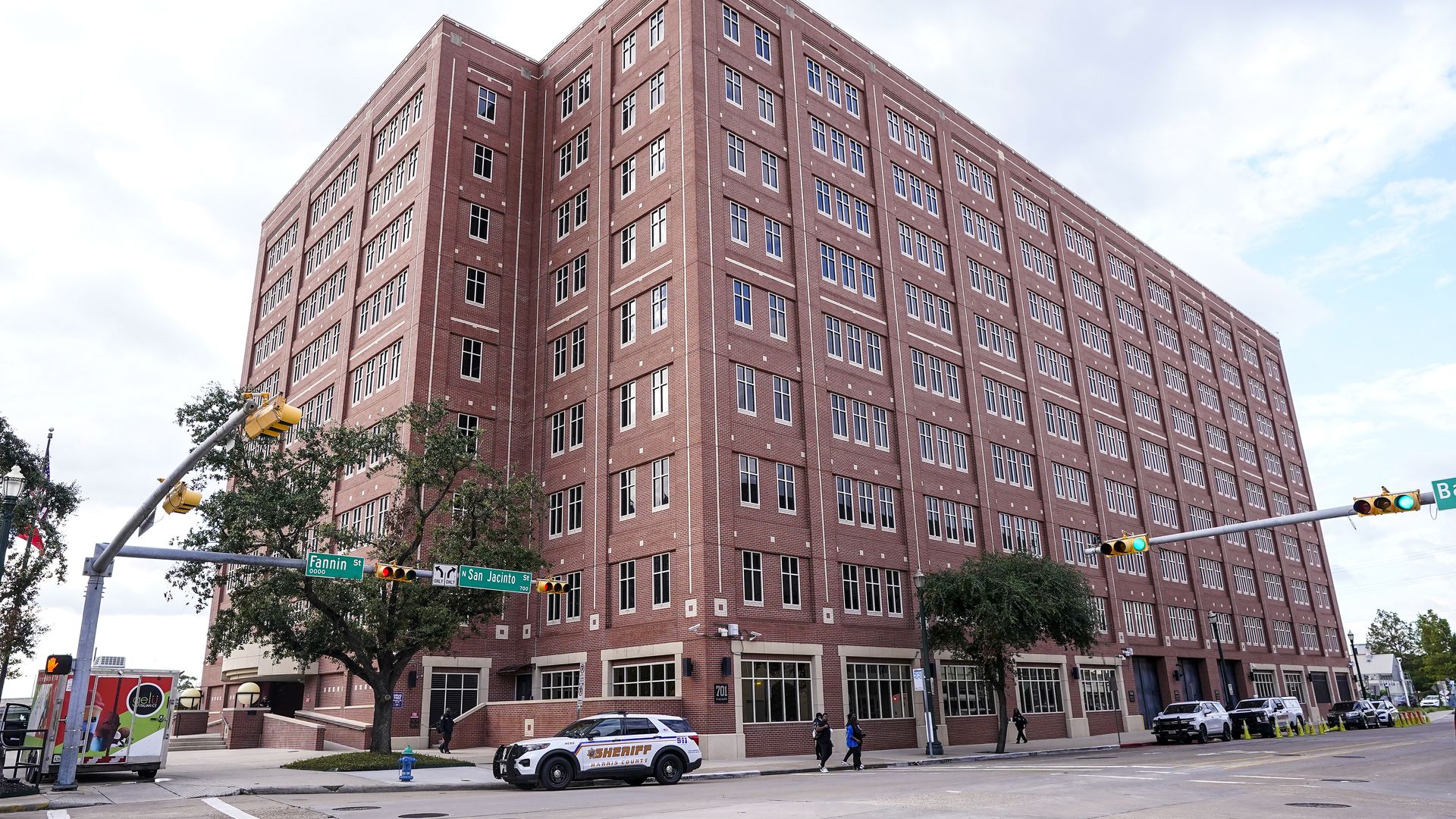 Harris County Jail, the eight-story red brick building at the corner of Fannin St and San Jacinto St, with traffic lights, a sheriff SUV, pedestrians, and cloudy sky.