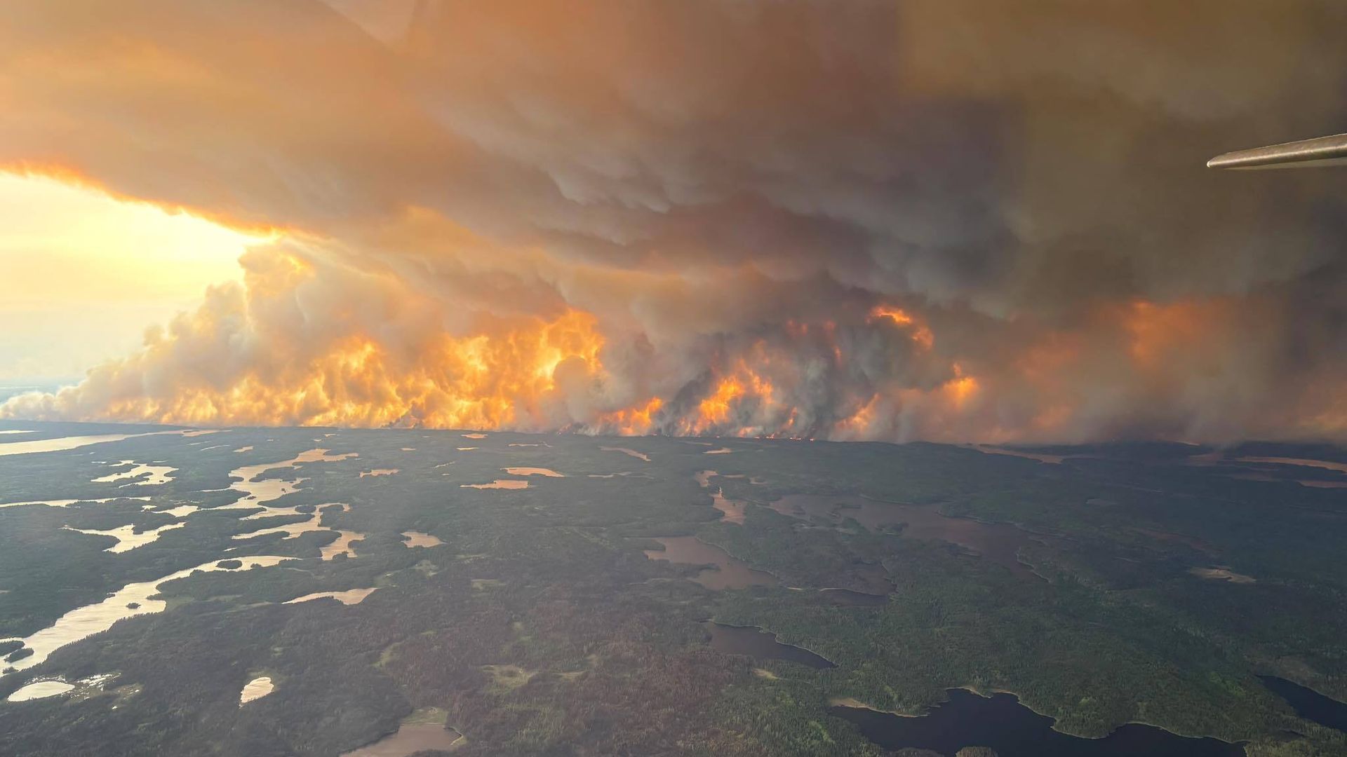 A large wildfire spreads across a forested landscape with lakes, producing thick, dark smoke and bright orange flames under a cloudy sky at sunset.