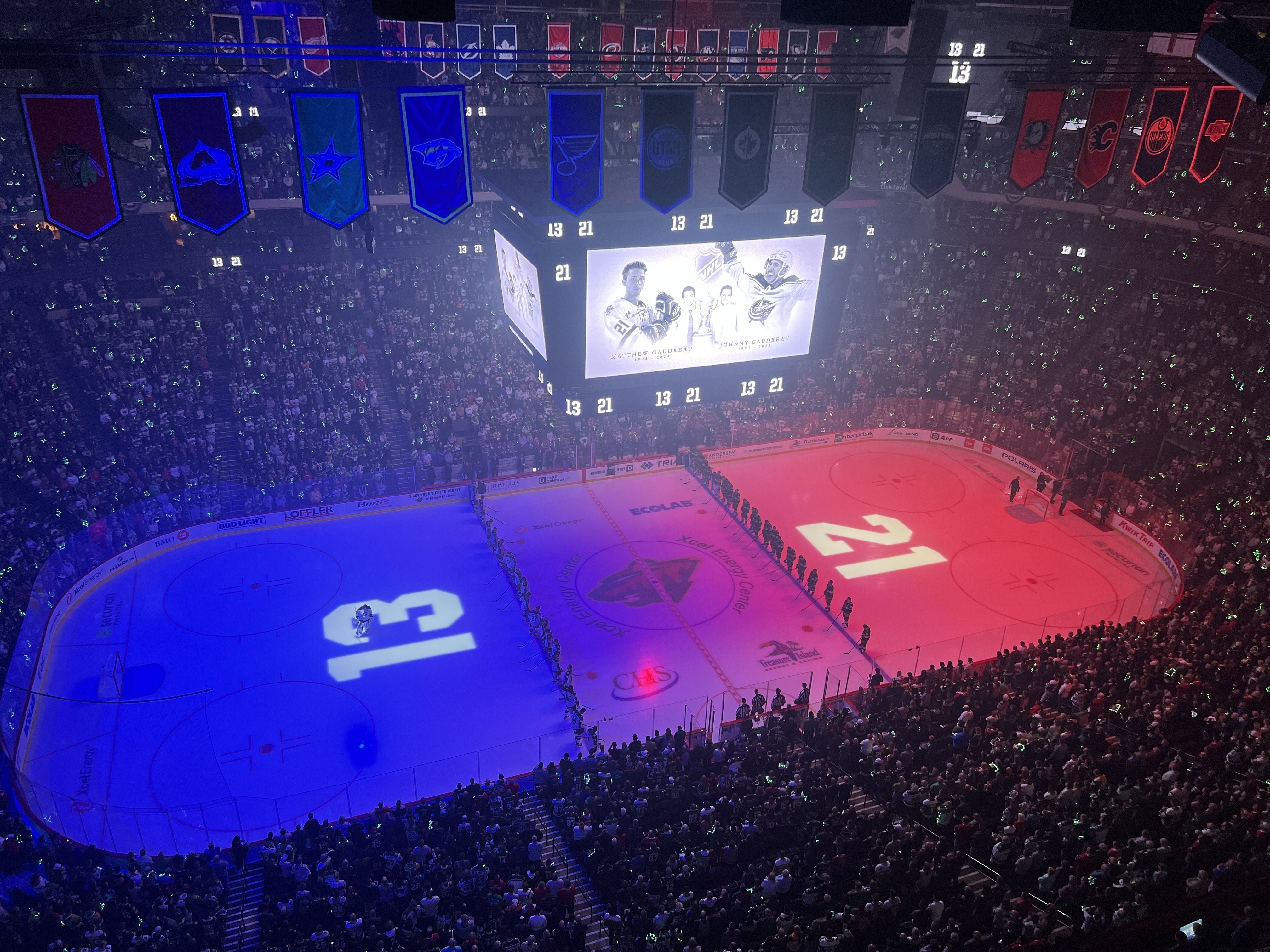 An overhead image of a darkened hockey rink bathed in blue and red light with the numbers 13 and 21 projected on the ice