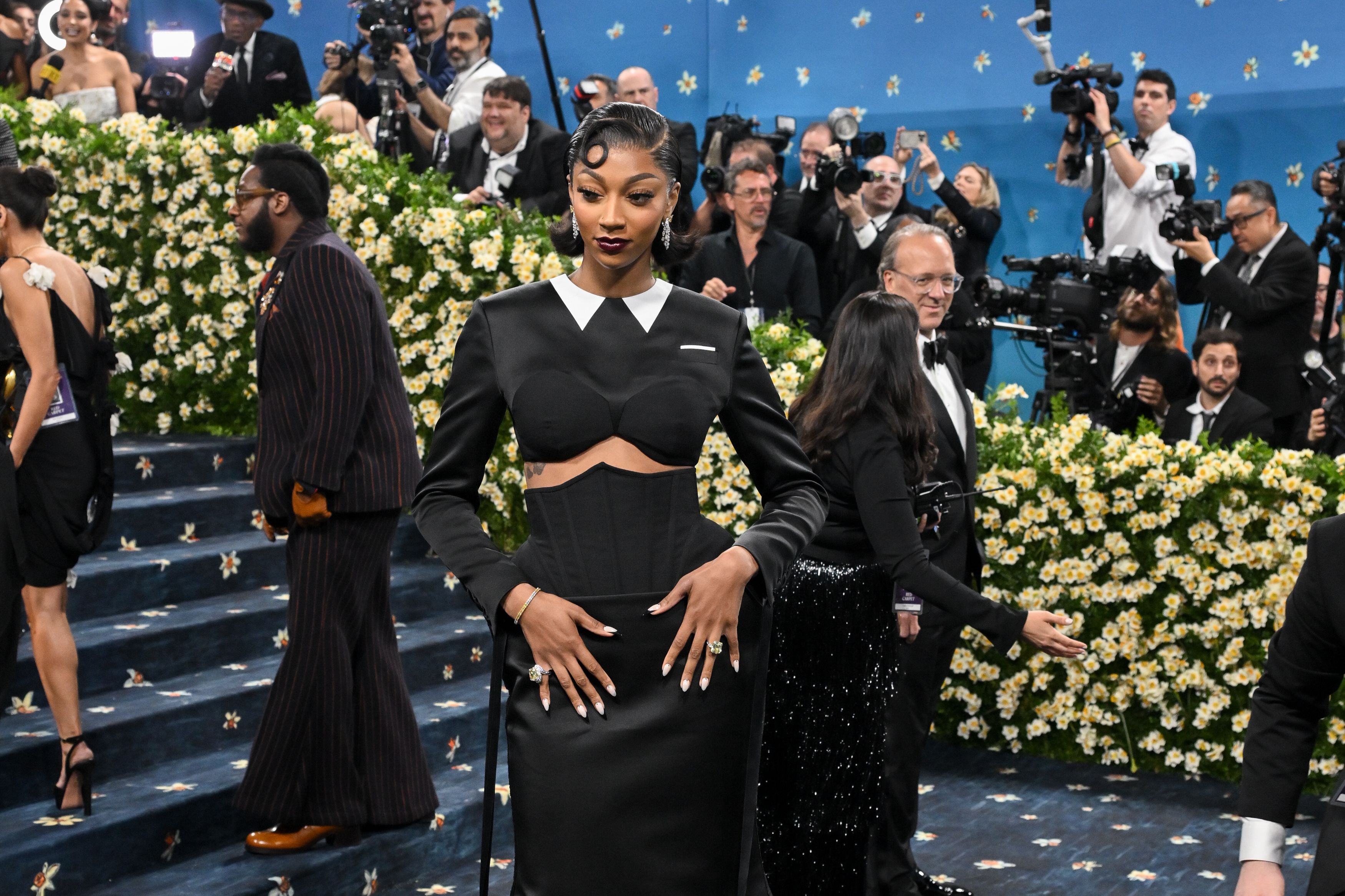 A glamorous woman in a black satin gown with a white collar poses on a blue carpet lined with white flowers as a crowd of photographers snaps pictures in the background.